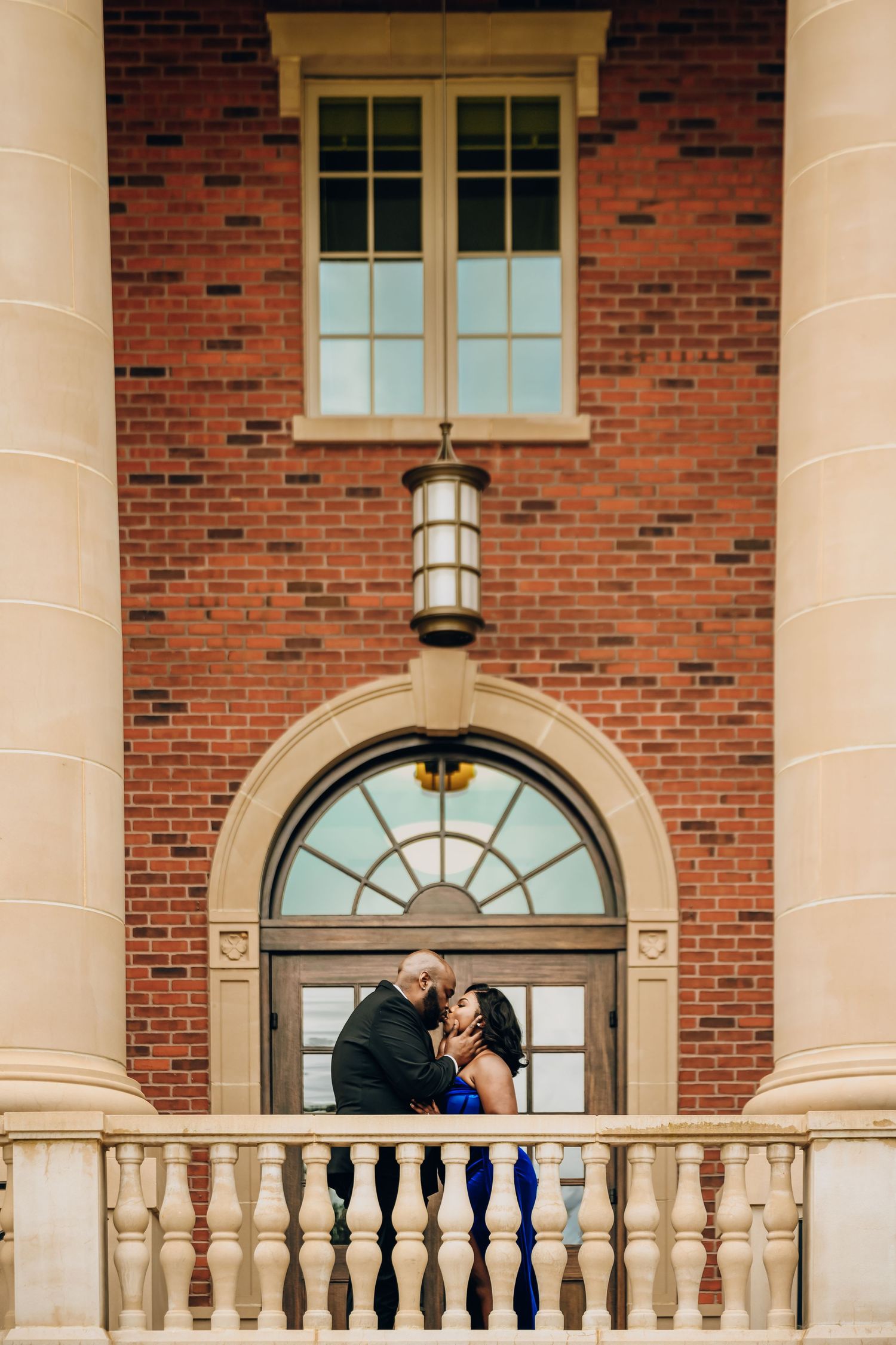 Couple shares romantic moment on brick building balcony with white columns.