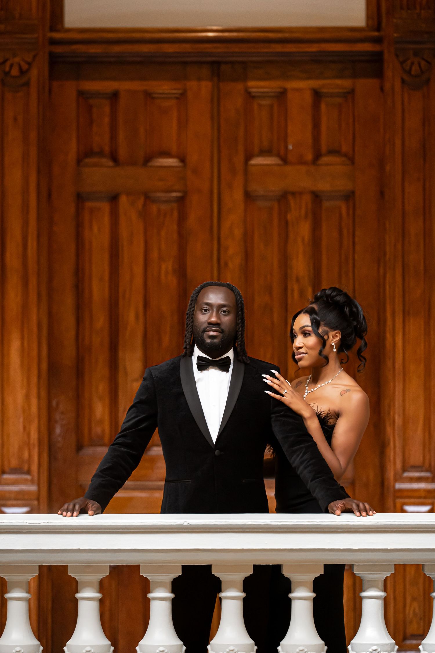 A couple stands together on an ornate balcony with wooden paneling in formal wedding attire.