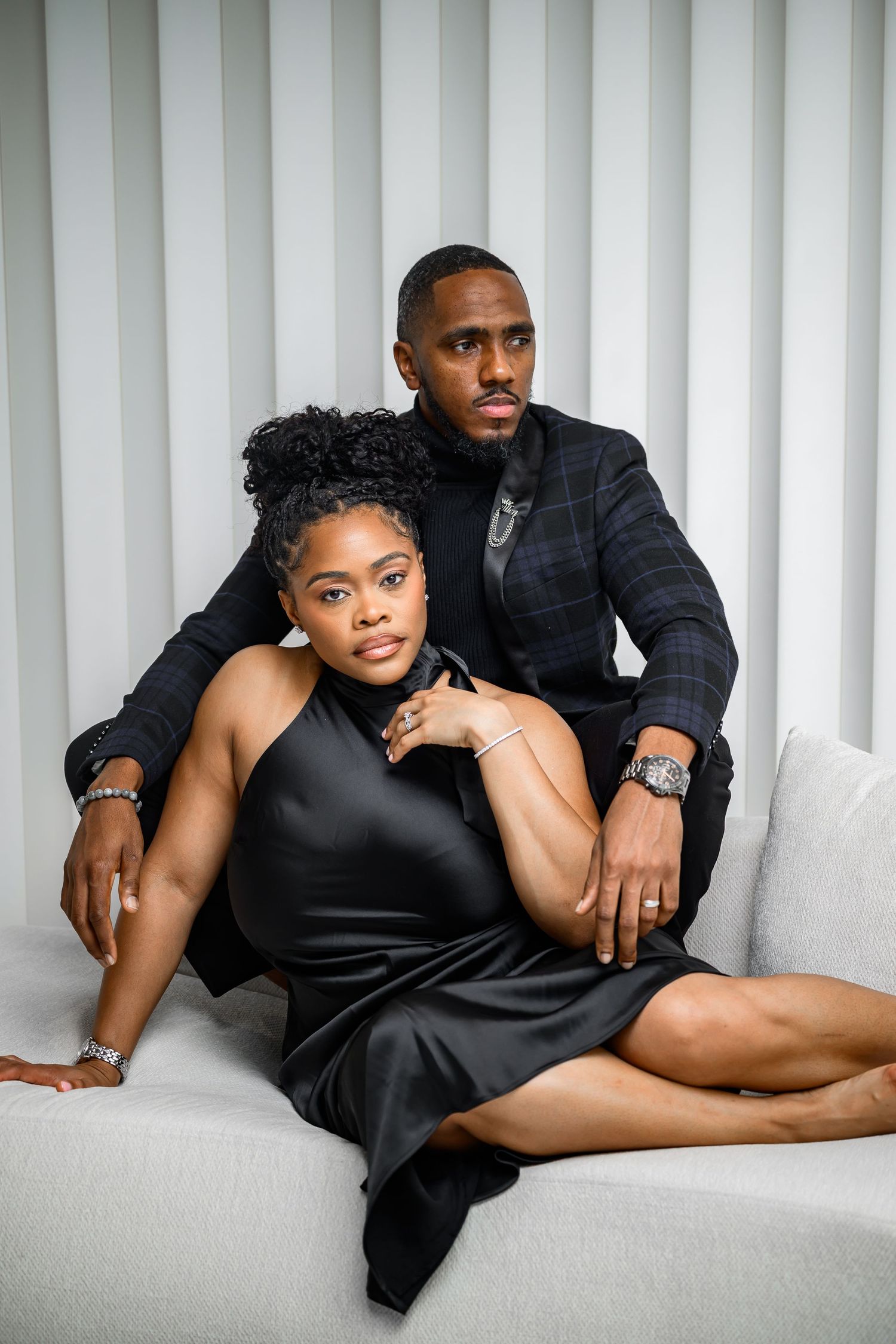 A couple poses intimately on a white couch in matching black formal attire.