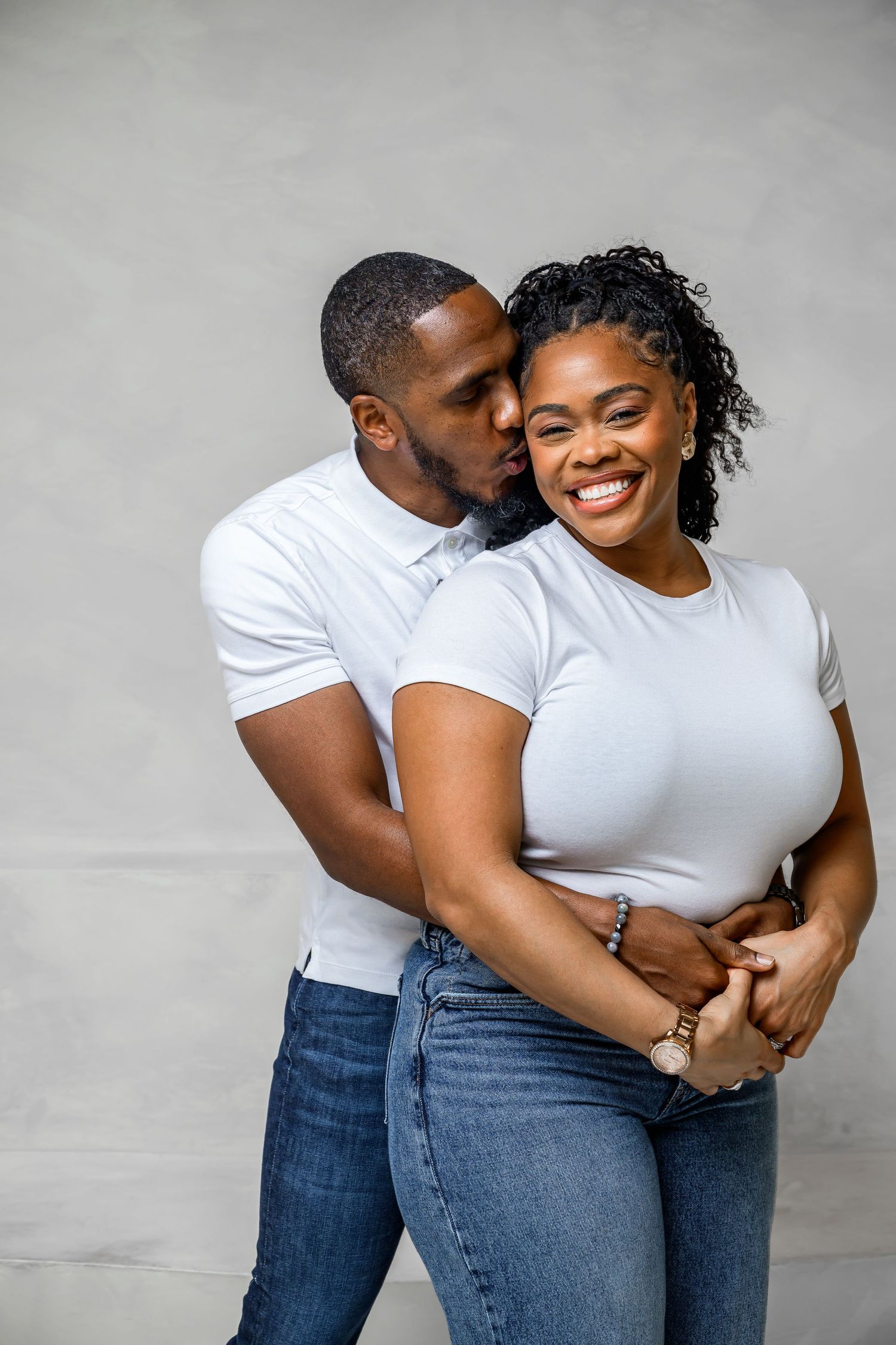 Affectionate couple in matching white t-shirts and jeans embrace and smile against a gray wall.