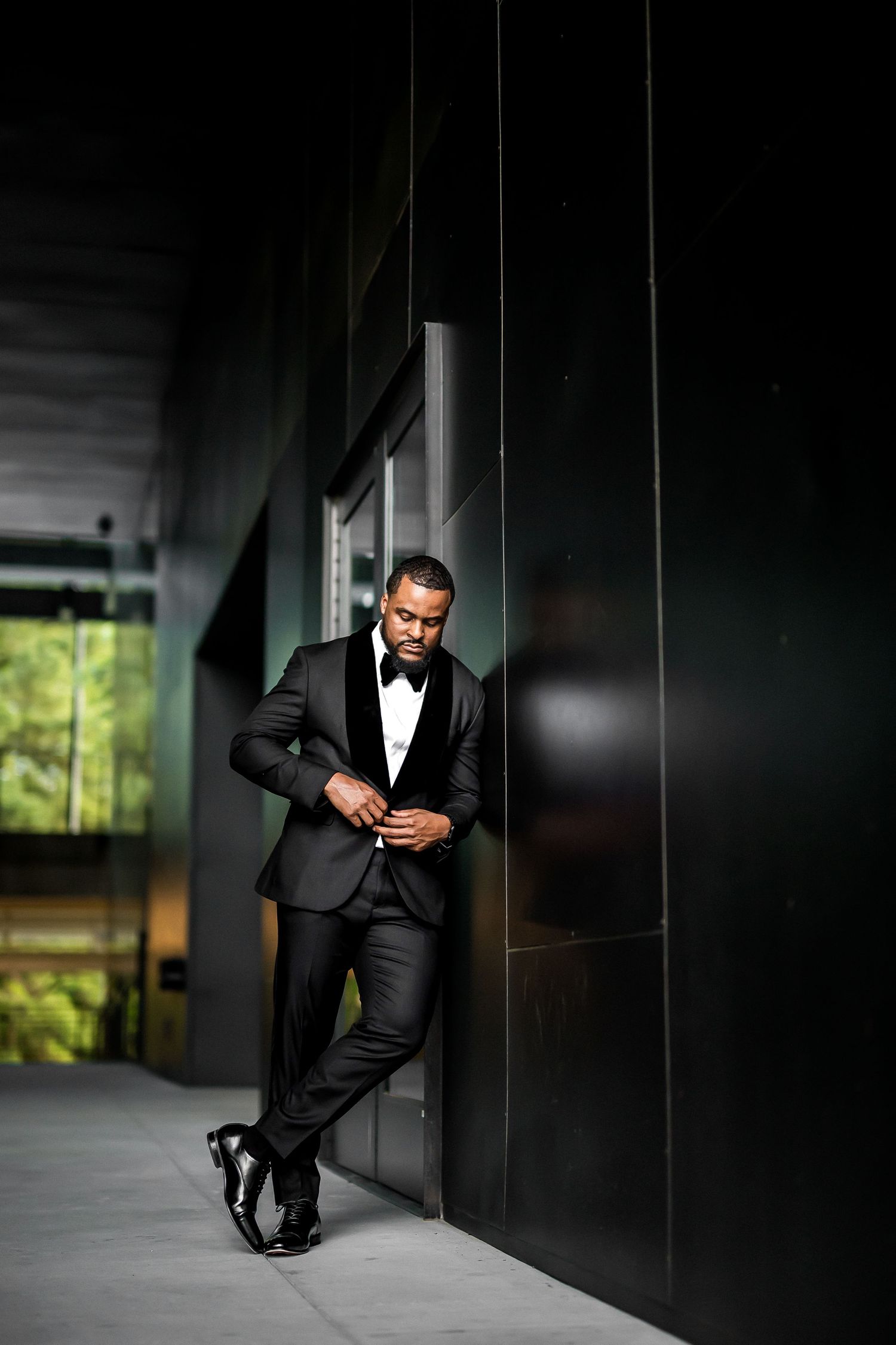 A well-dressed figure in a dark tuxedo and bowtie leans against a modern black wall in dramatic lighting.
