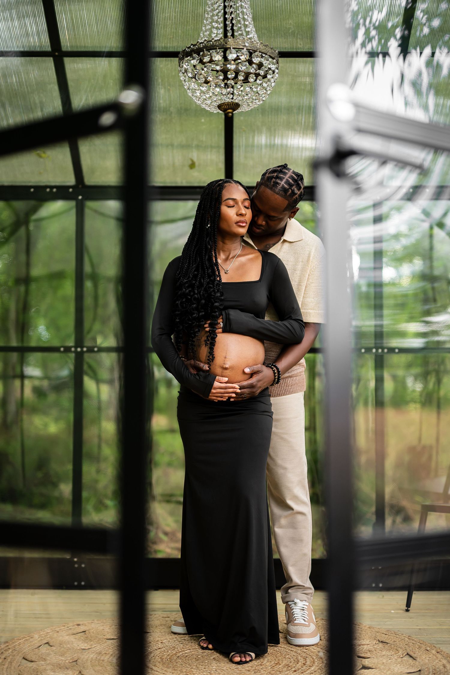 Intimate maternity photoshoot in a sunlit greenhouse with elegant crystal chandelier overhead.