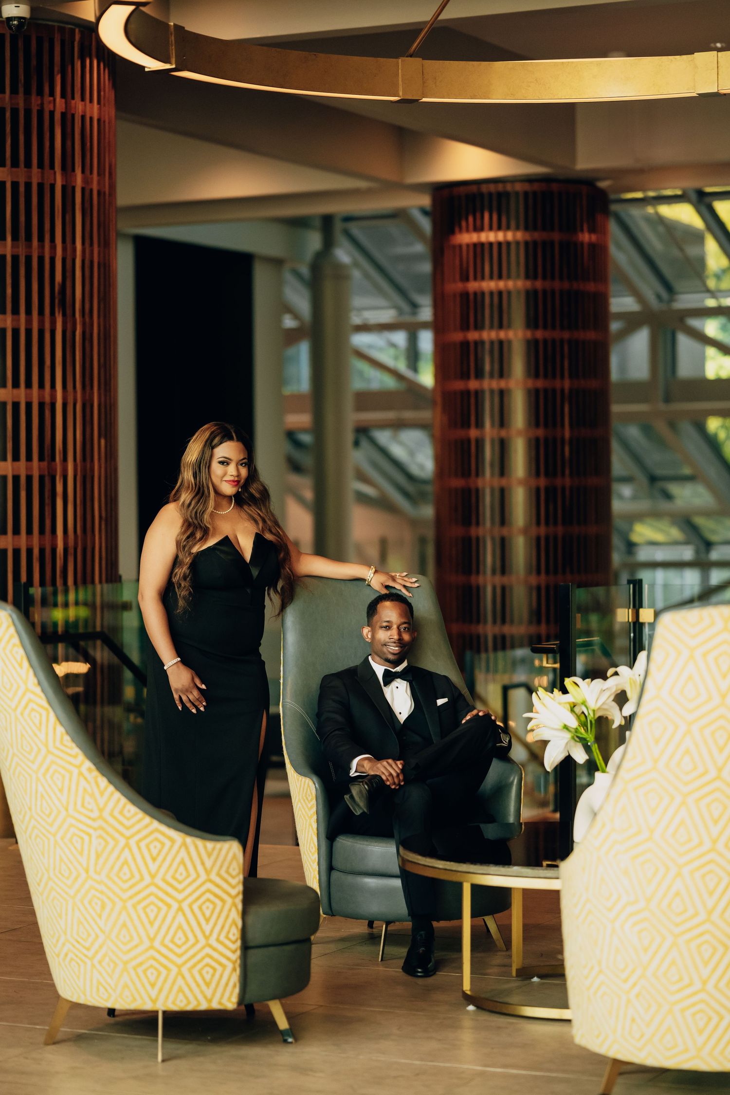 Couple in formal attire posing on stylish chairs in modern hotel lobby.