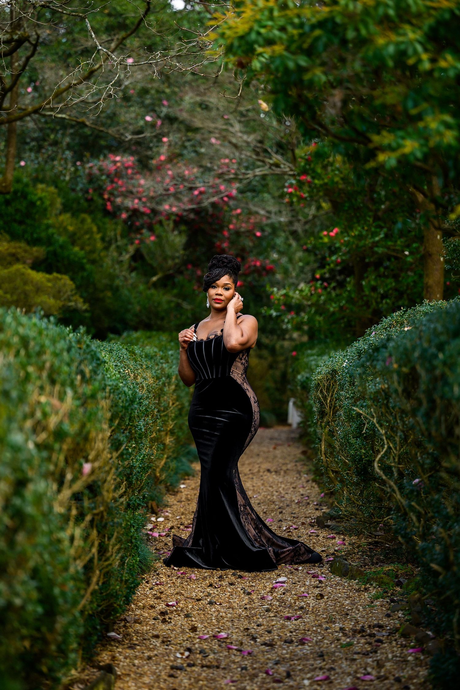 A figure in a black dress sits contemplatively on a winding garden path surrounded by lush greenery and flowers.