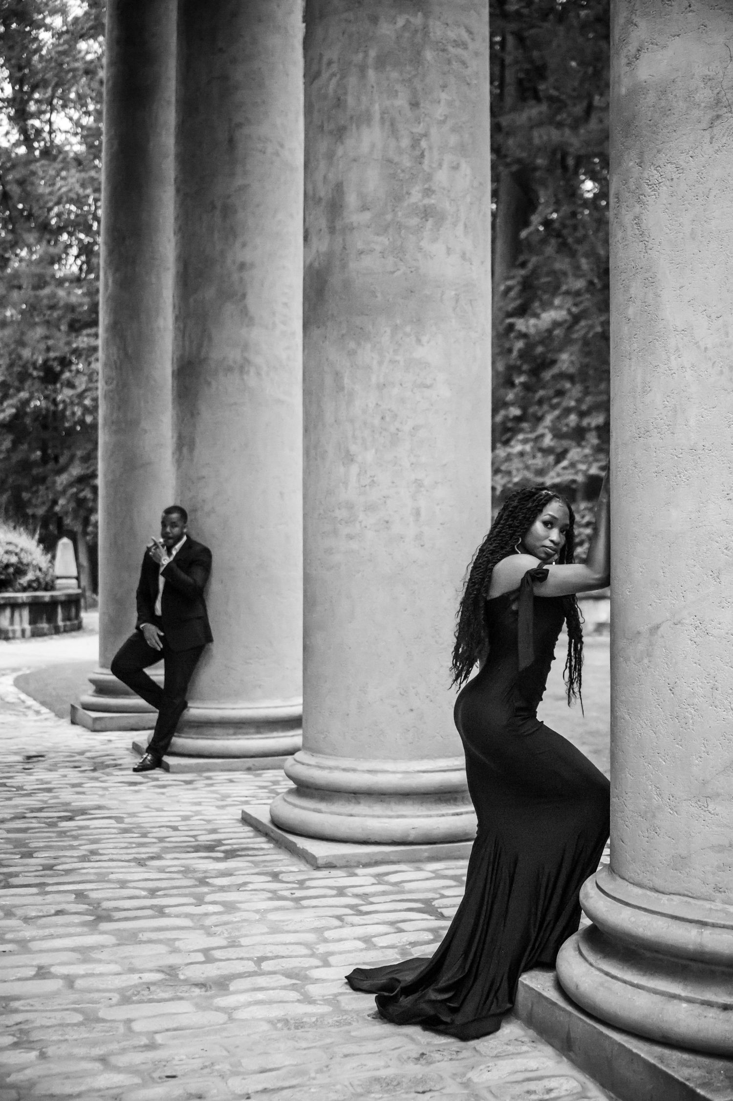 Elegant couple in formal black attire pose dramatically against classical stone columns in black and white photography.