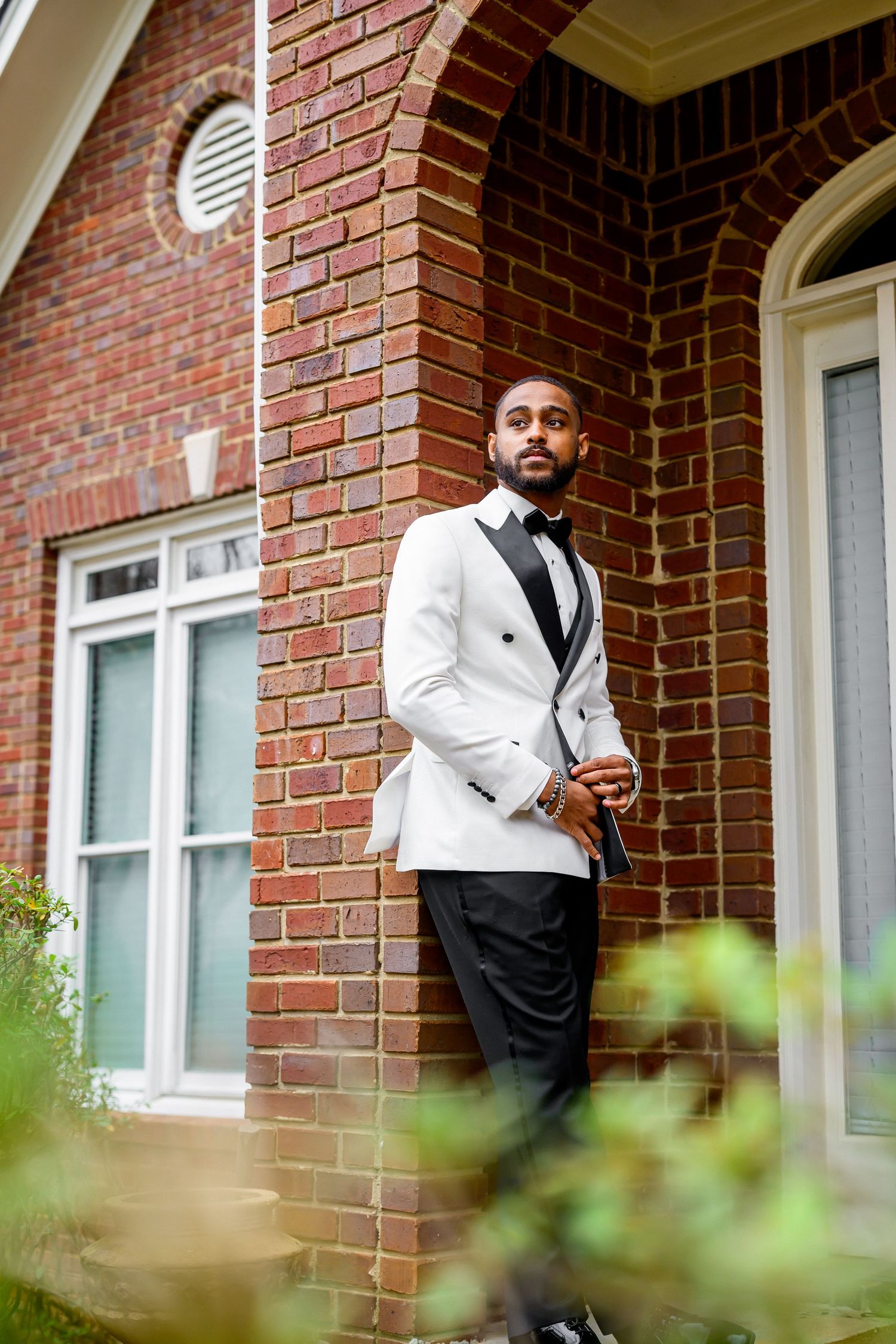 A person in a white dinner jacket and black vest poses against a brick wall exterior with windows.