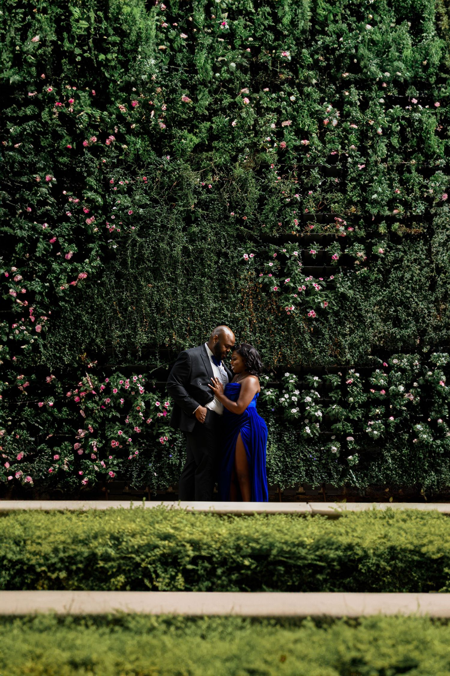 Couple poses against lush green hedge wall in outdoor garden setting.