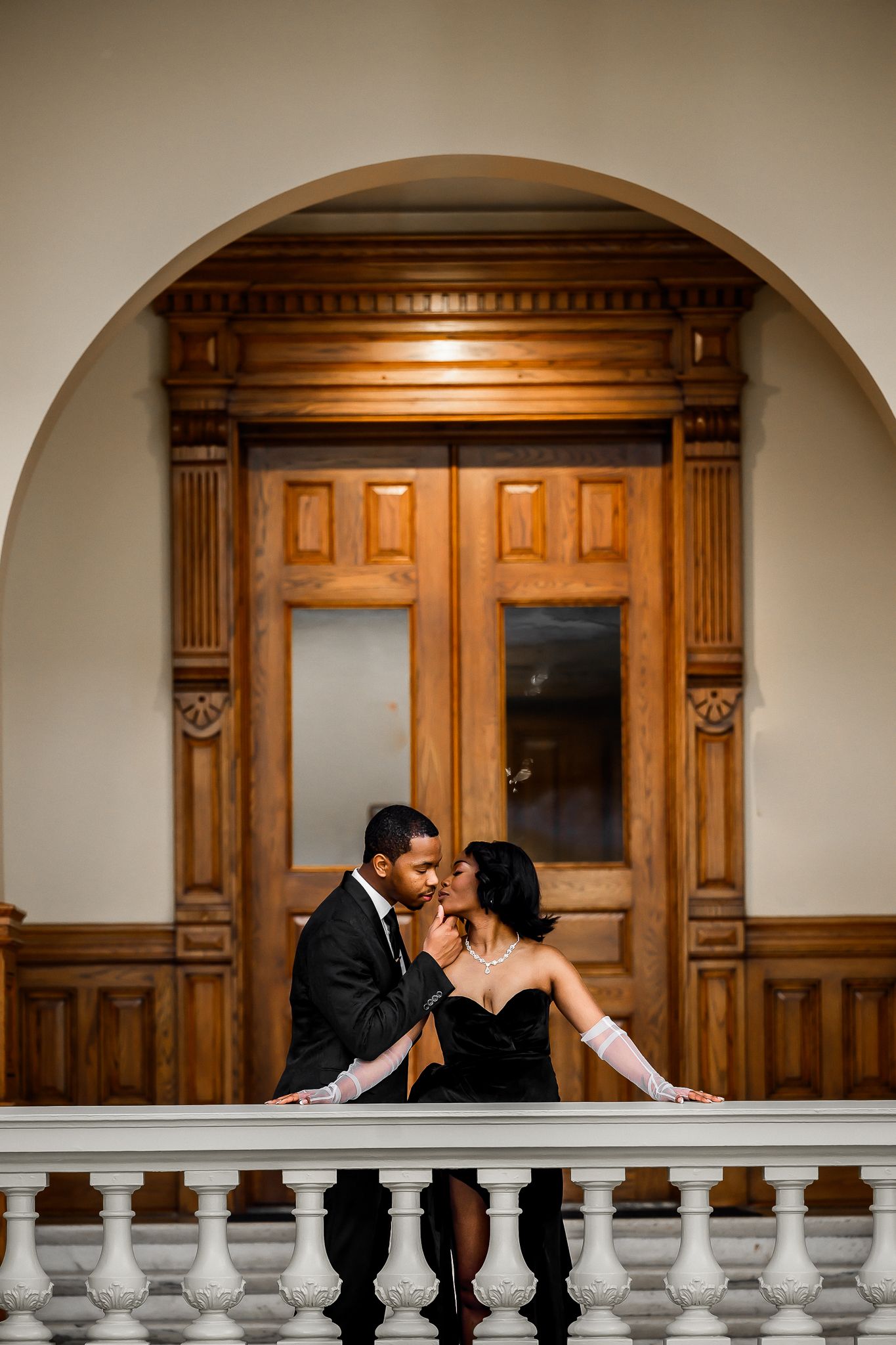 Romantic couple in formal wear share intimate moment by ornate wooden doorway and balustrade.