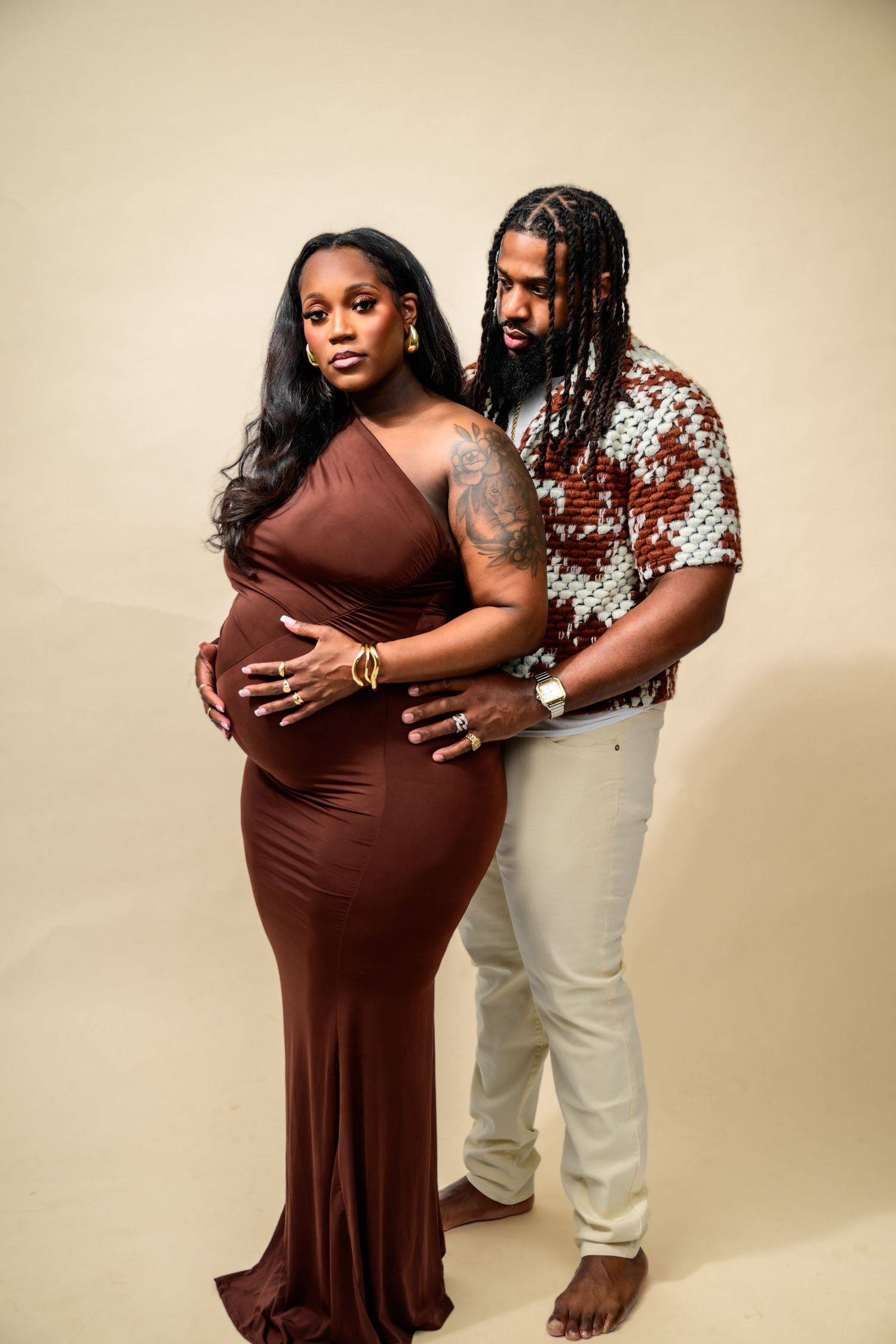 Loving maternity poses in brown dress with partner against neutral background.