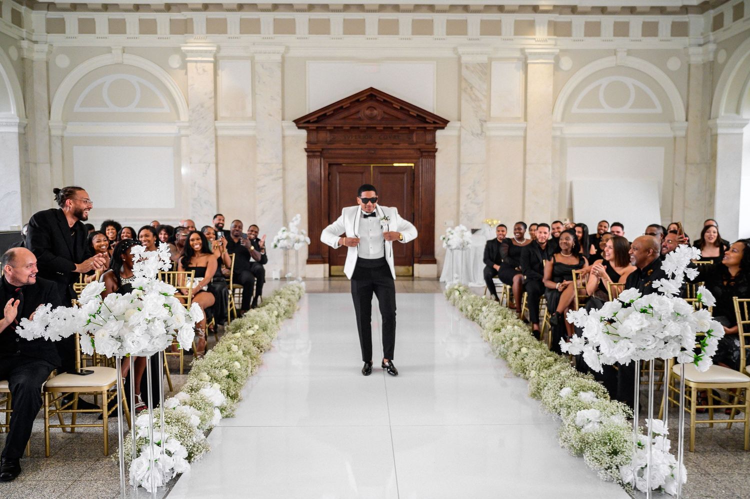 Wedding ceremony setup in an elegant white venue with rows of chairs and floral arrangements along the aisle.