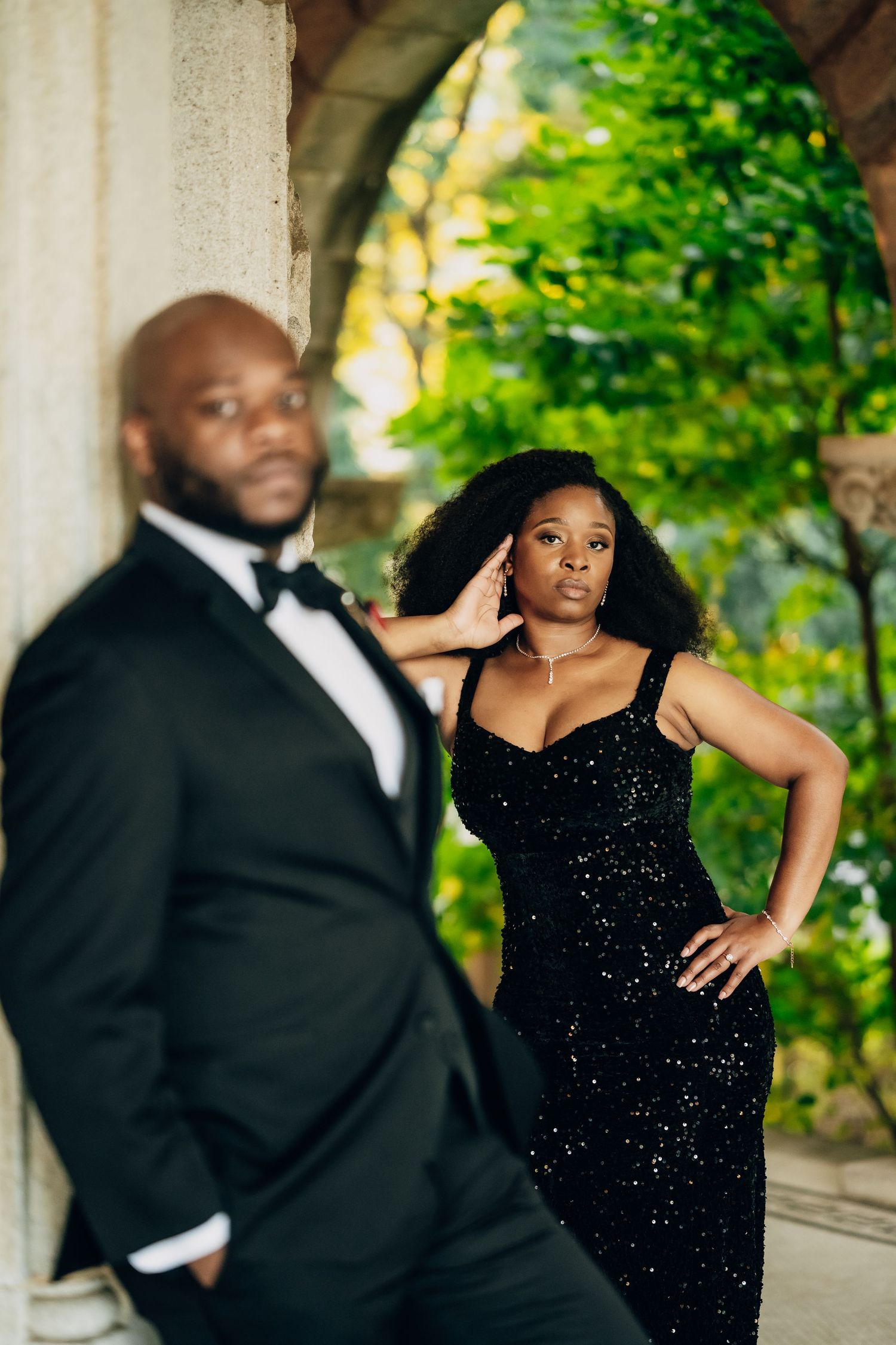 Couple in formal wear poses dramatically against stone columns and green foliage.