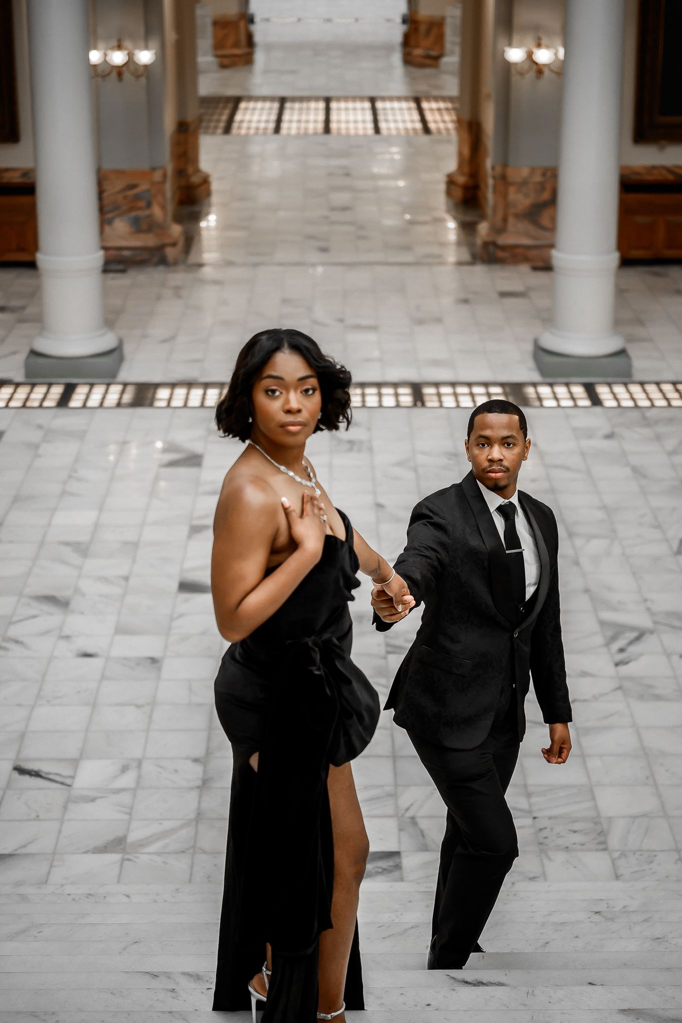 Couple in black formal wear posing dramatically on marble floor with classical architecture.