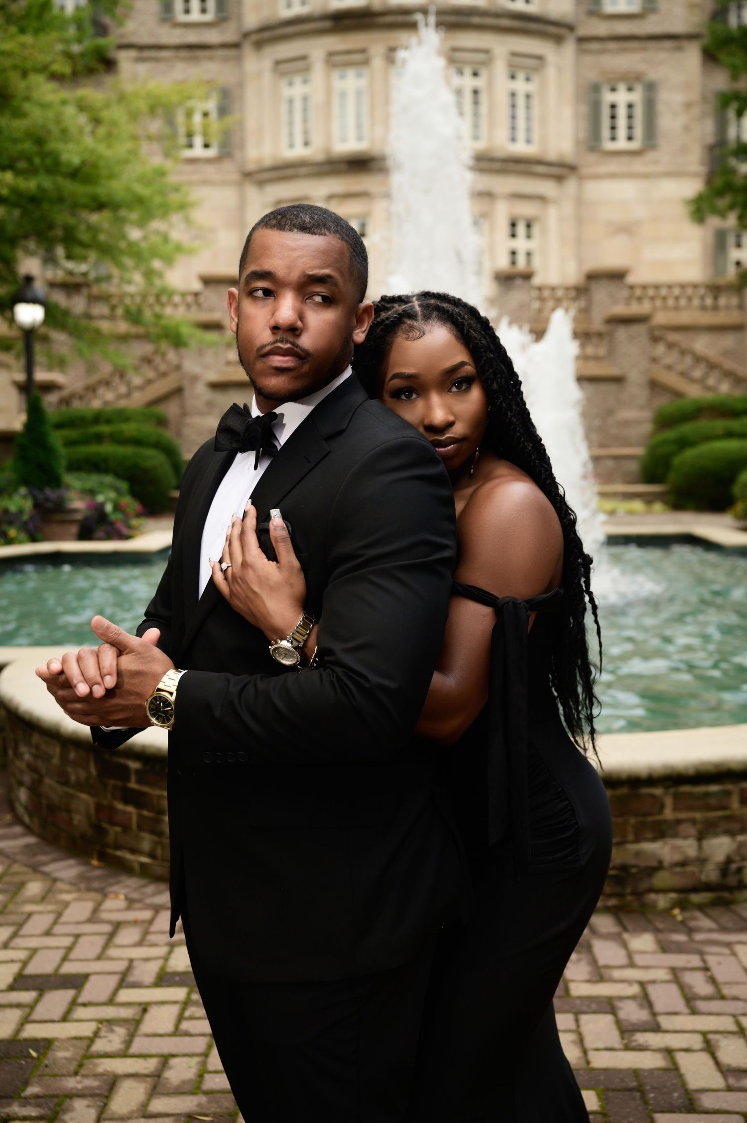 Romantic couple poses by a stone fountain in formal attire.