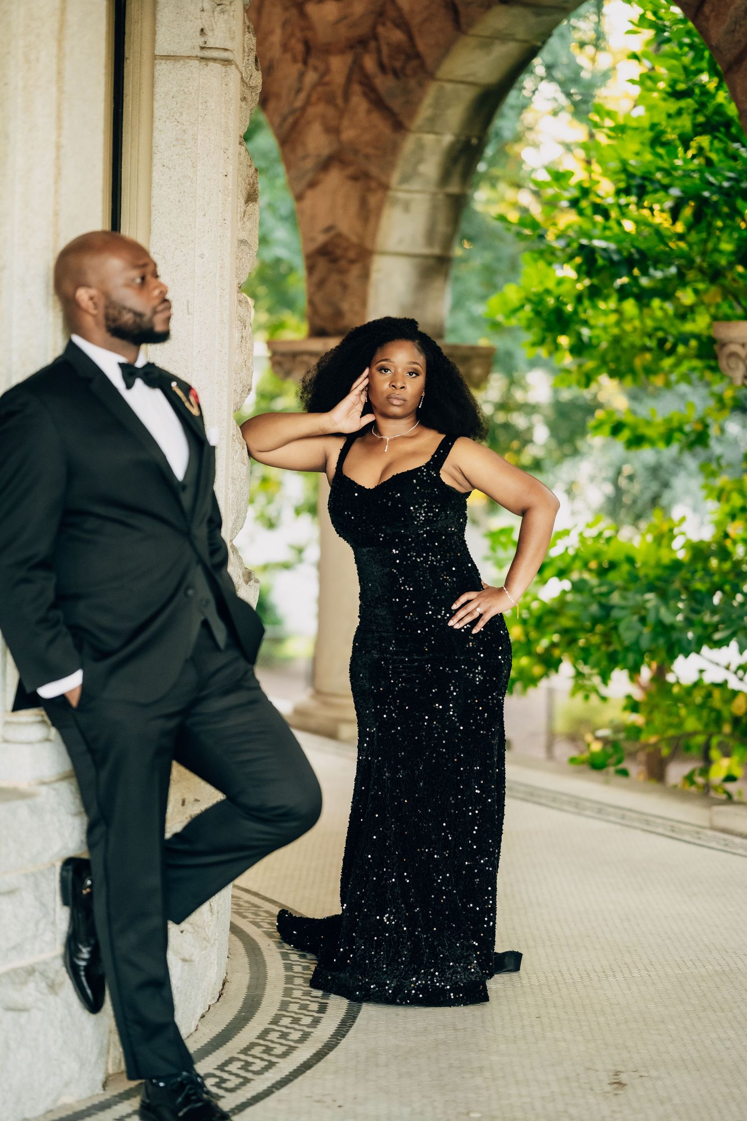 Playful couple in formal attire poses against stone columns and greenery.