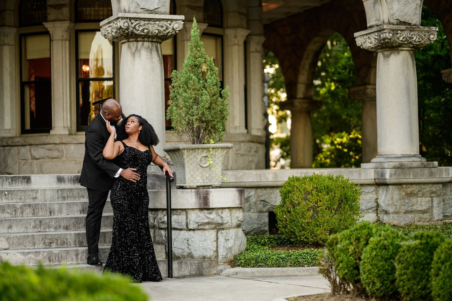 Couple in formal attire embrace on stone steps surrounded by classic architecture and greenery.