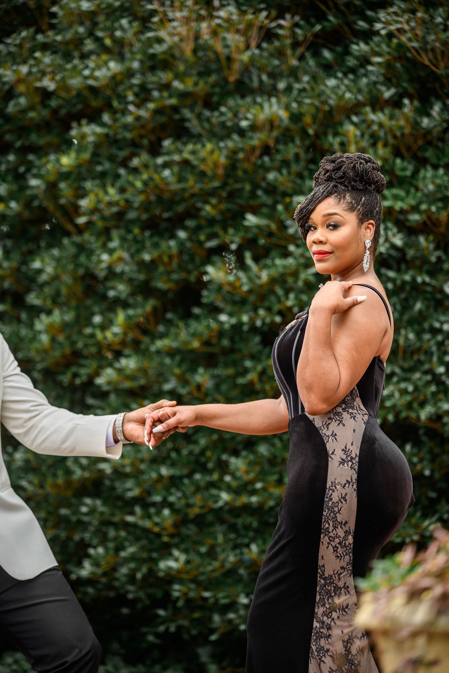 Elegant couple in formal attire holding hands against lush green garden backdrop during romantic photoshoot.
