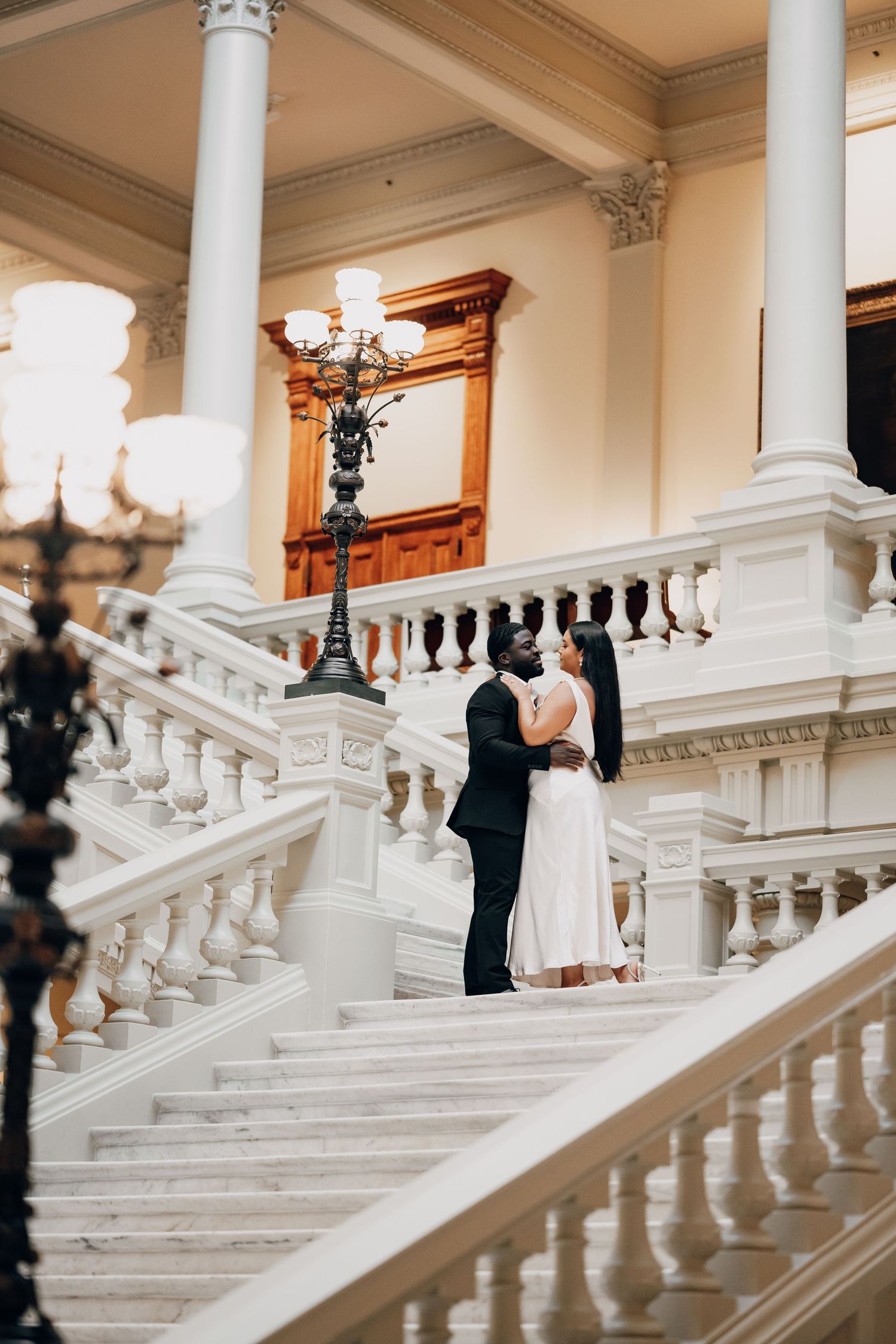 Couple in formal attire standing on grand white marble staircase during wedding photoshoot.