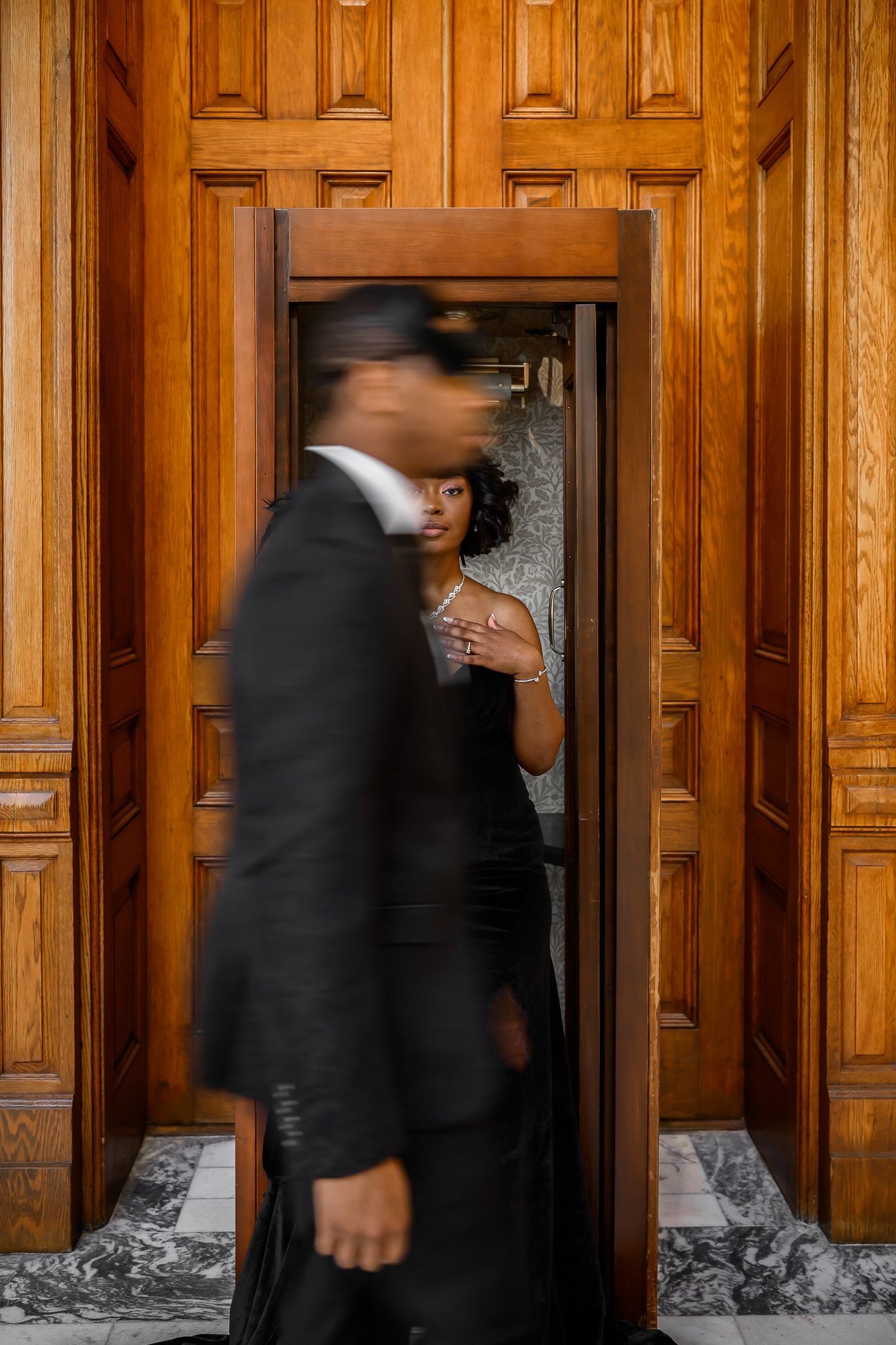 People in business attire walk past wooden paneled walls with ornate architectural details in a formal building hallway.