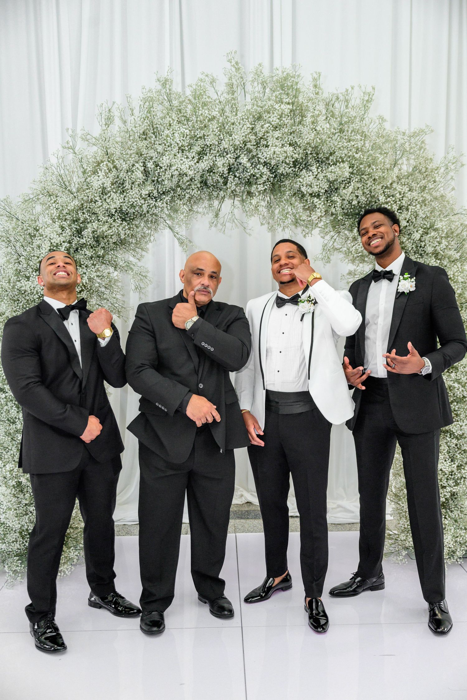 Groomsmen in black tuxedos pose playfully in a row against a white backdrop with greenery.