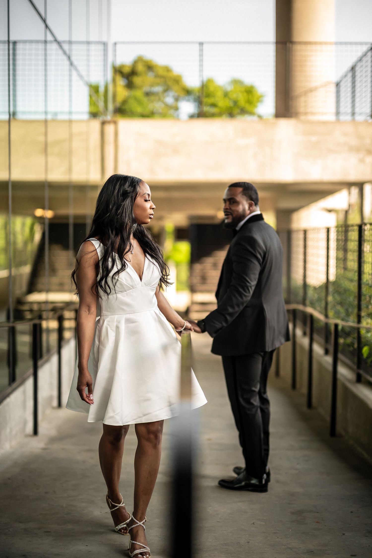 A couple in elegant formal wear poses on a modern concrete walkway with natural lighting.