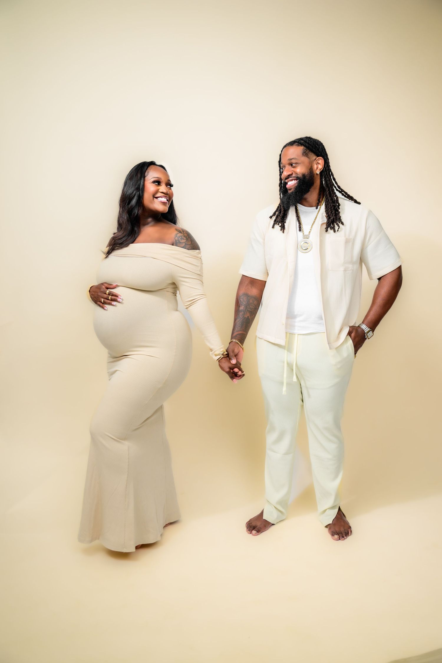 Studio maternity photoshoot with couple in white and cream outfits against neutral background.
