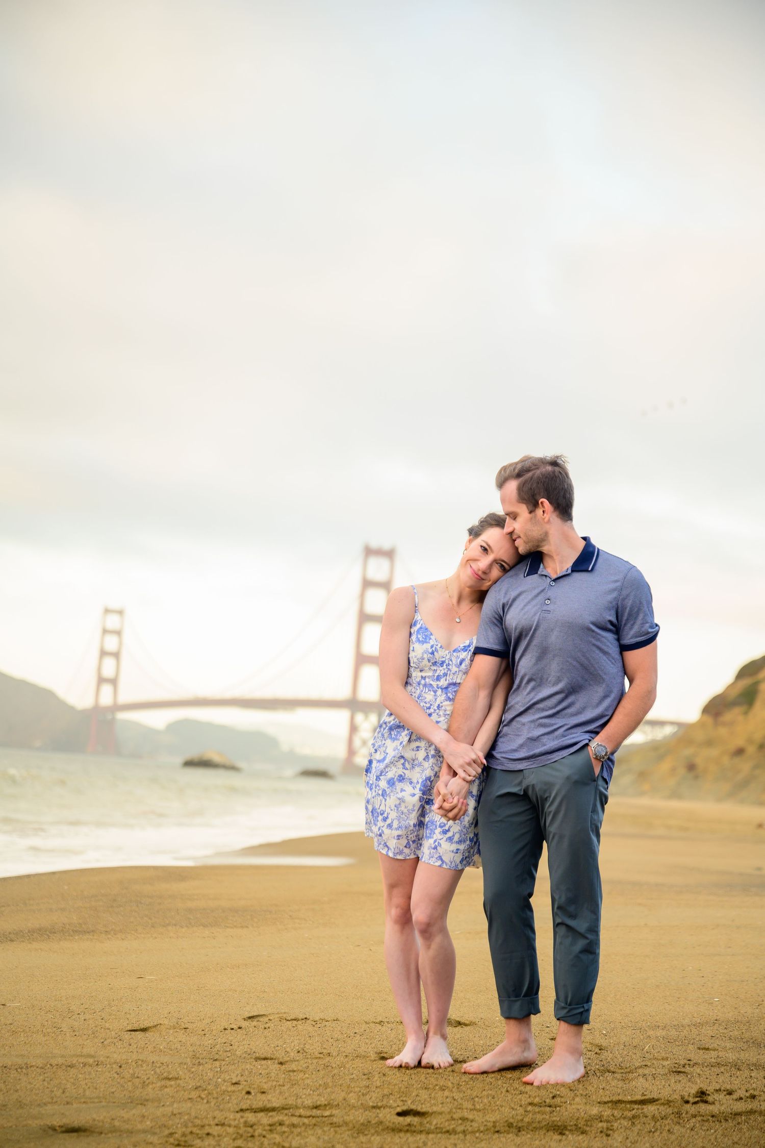 A couple poses on Baker Beach with the Golden Gate Bridge visible in the background.