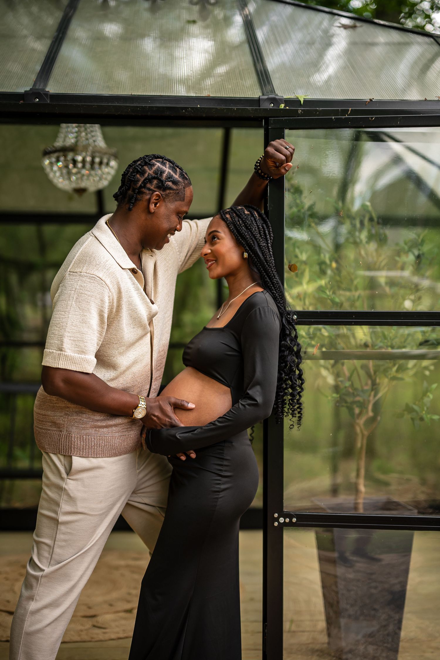 An intimate maternity photoshoot features a couple in a sunlit greenhouse setting.