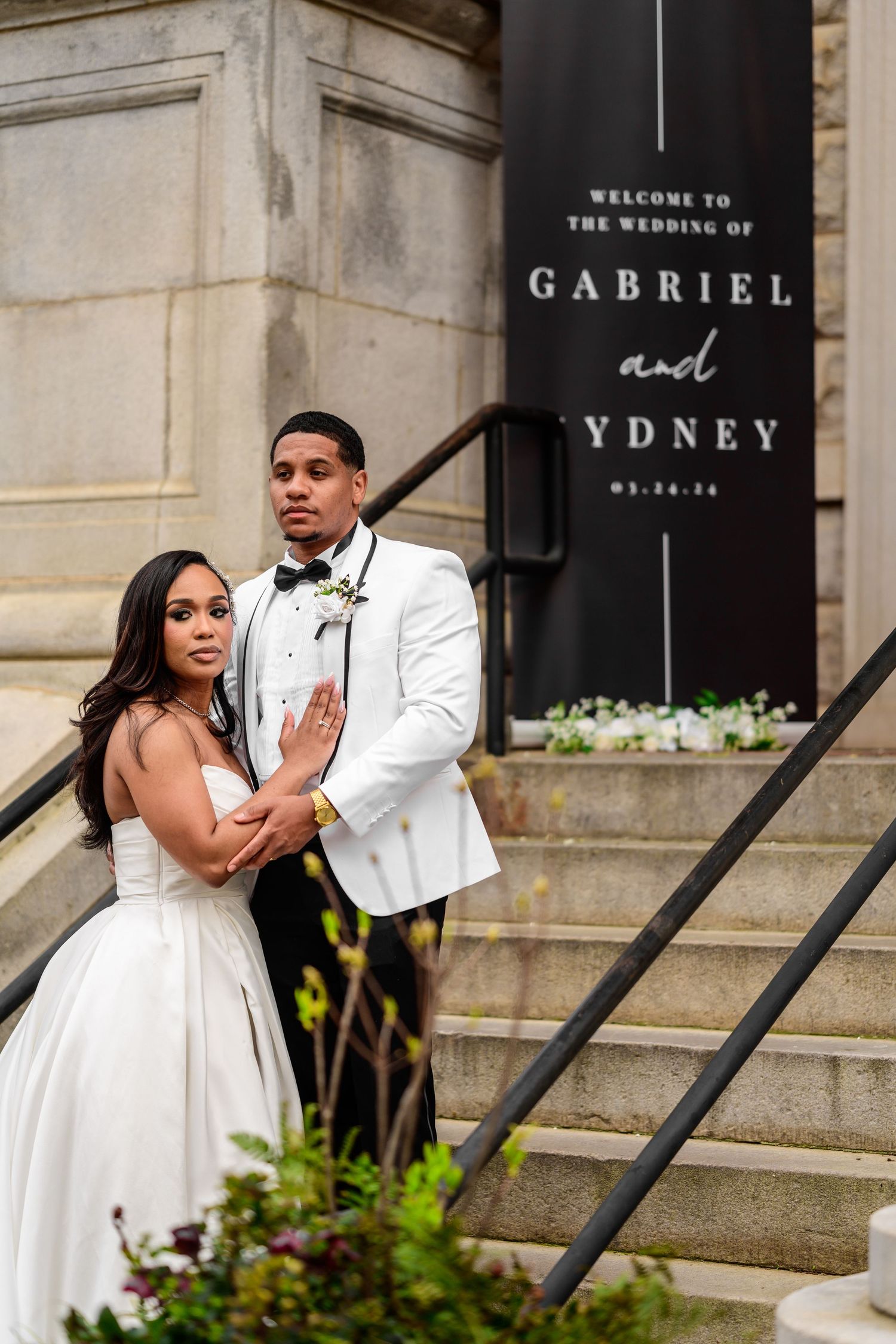 Wedding couple poses by stone steps with elegant black signage in background.