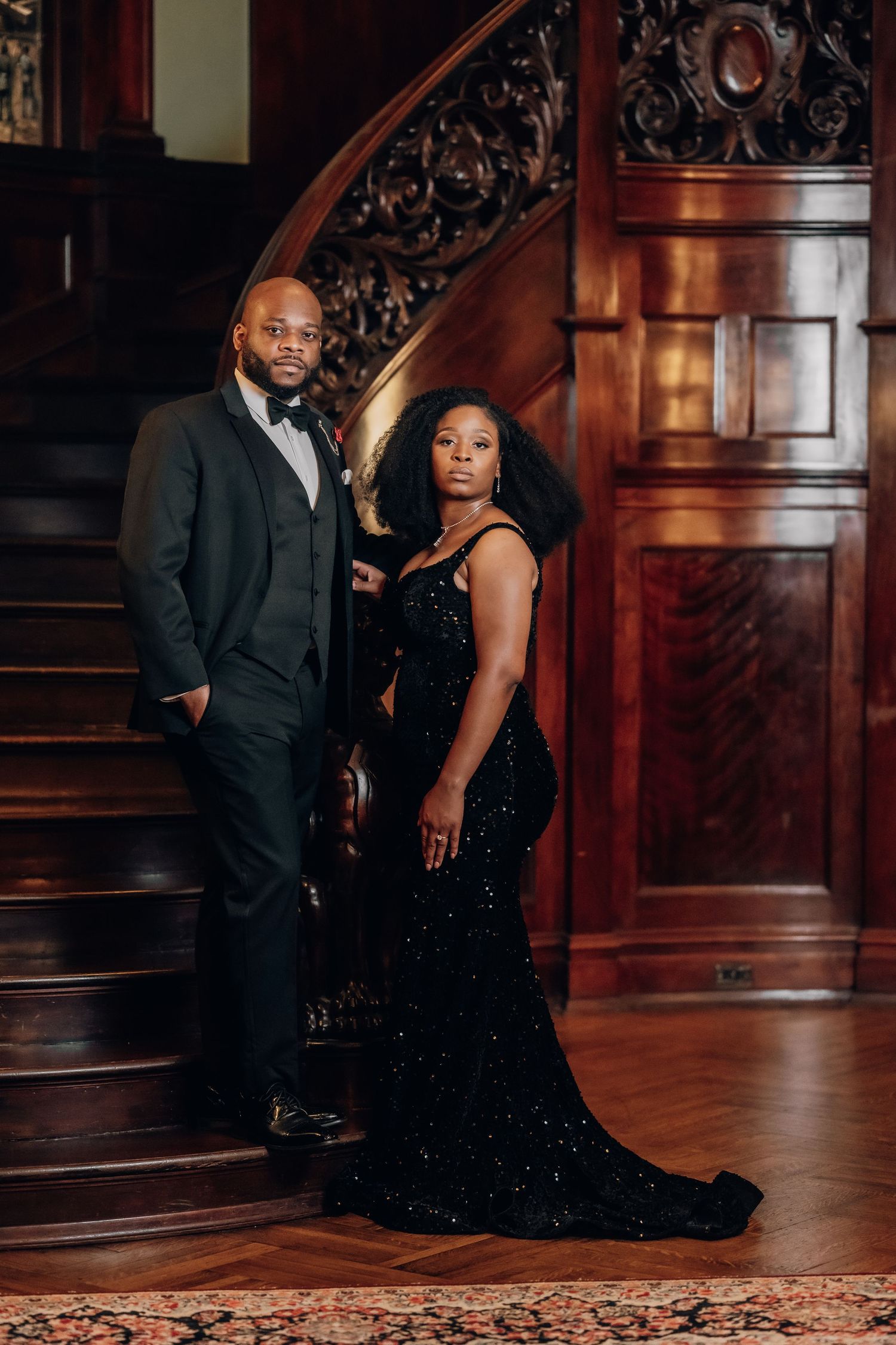 Elegant couple in formal attire poses on grand wooden staircase during romantic photoshoot.