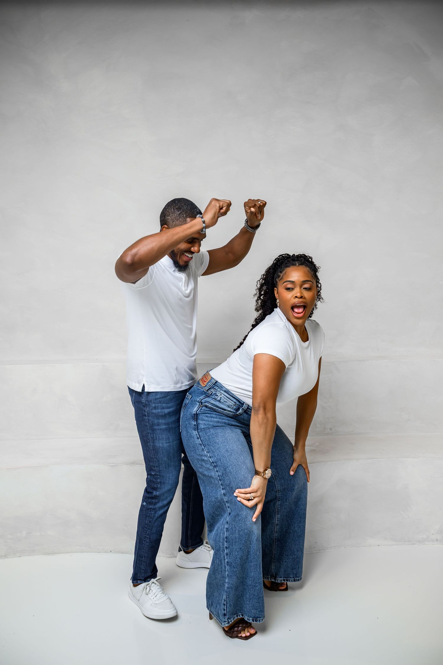 Two people in matching outfits of jeans and white shirts perform a playful dance move sequence against a gray backdrop.