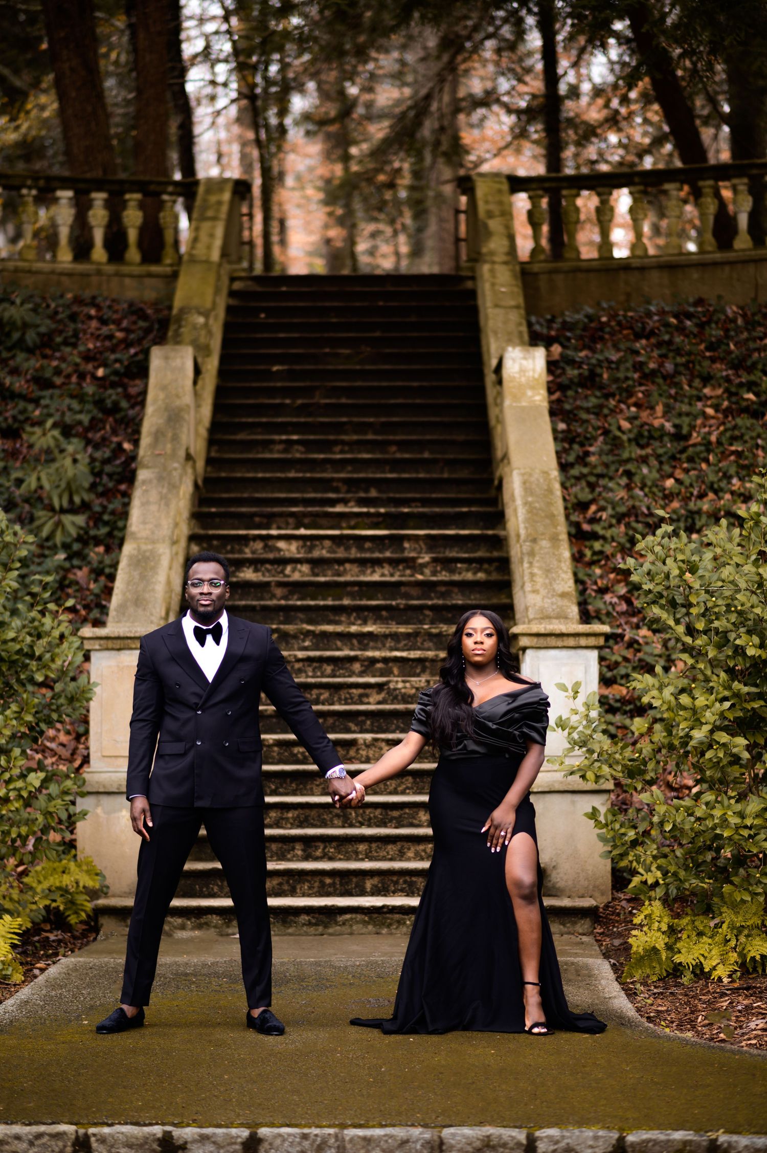 Glamorous couple in black formal wear pose on grand outdoor stone staircase.