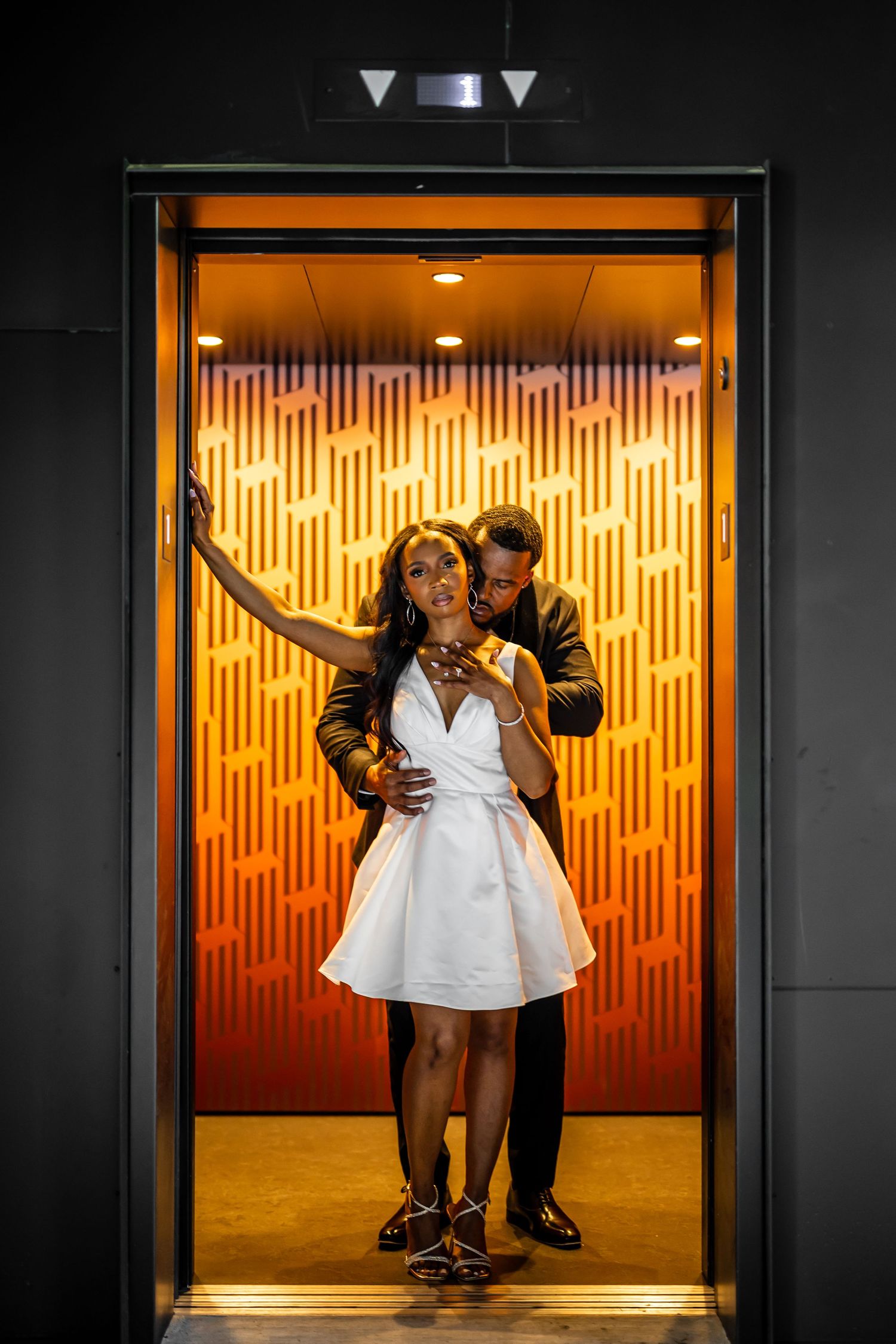 A stylish couple poses in an elevator with dramatic orange backlighting and geometric patterns.