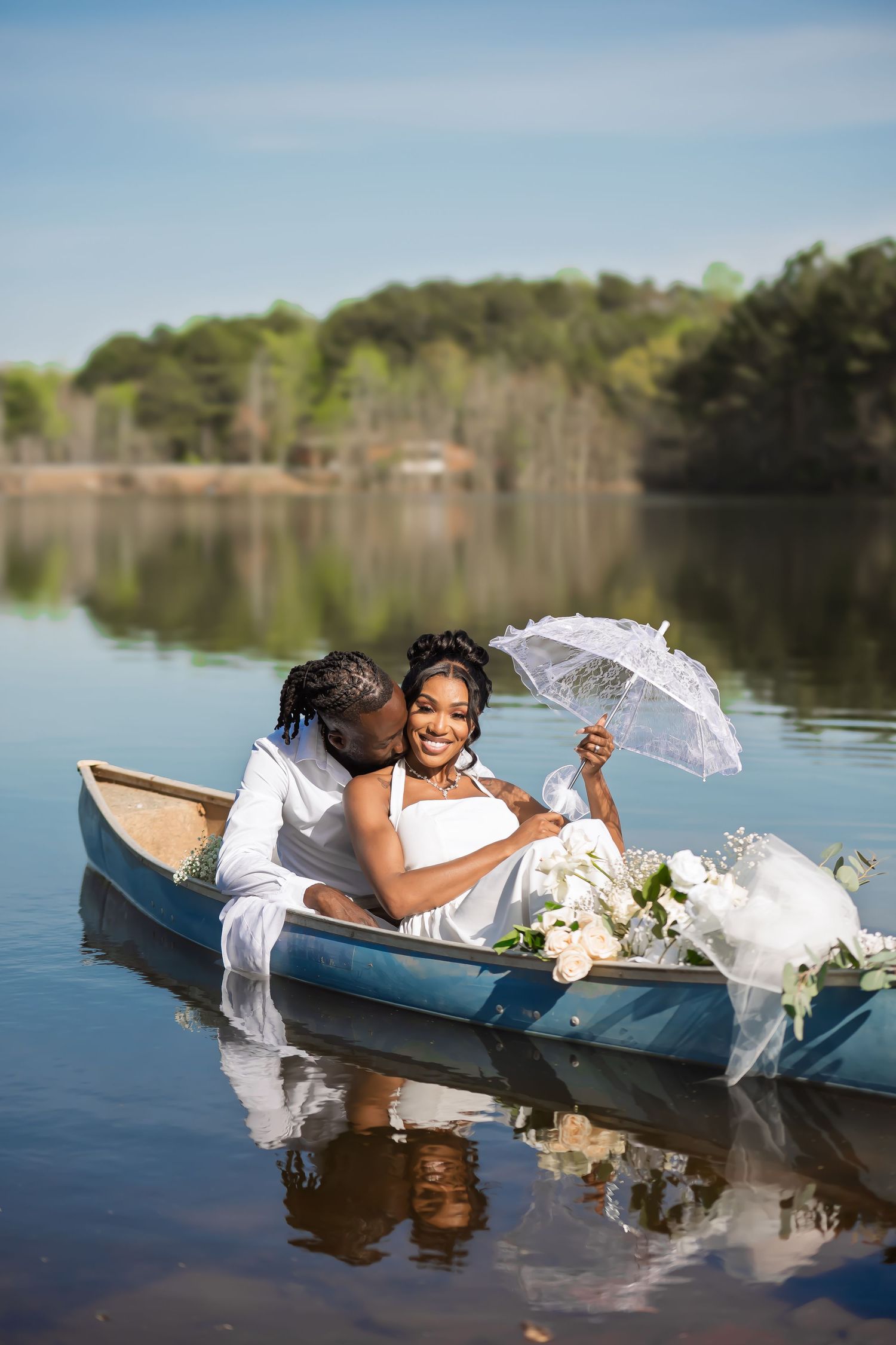 Romantic wedding photo of couple in canoe with white parasol and floral arrangements on calm lake.