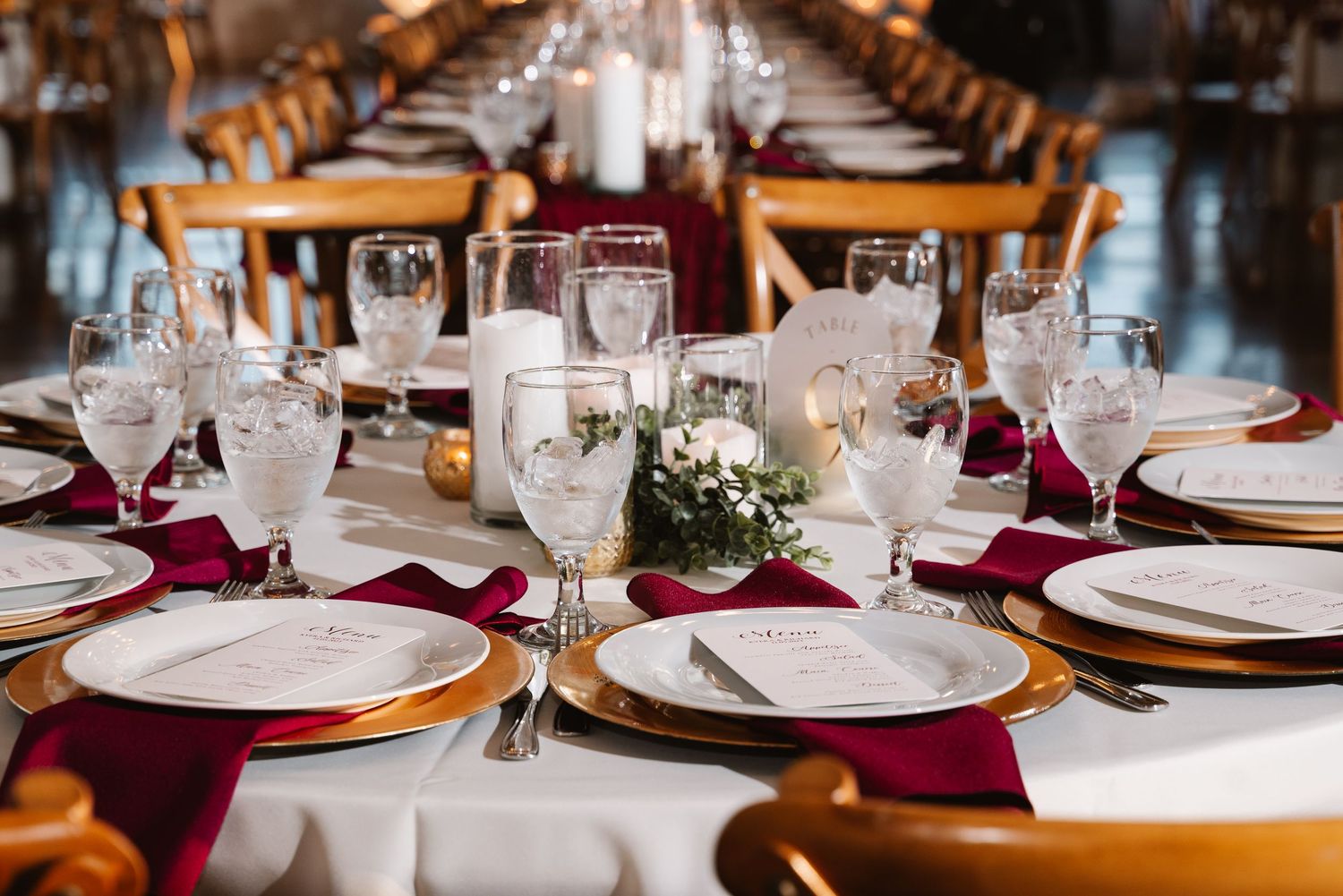 Elegant wedding reception tables set with burgundy napkins, white plates, and crystal glasses under warm lighting.