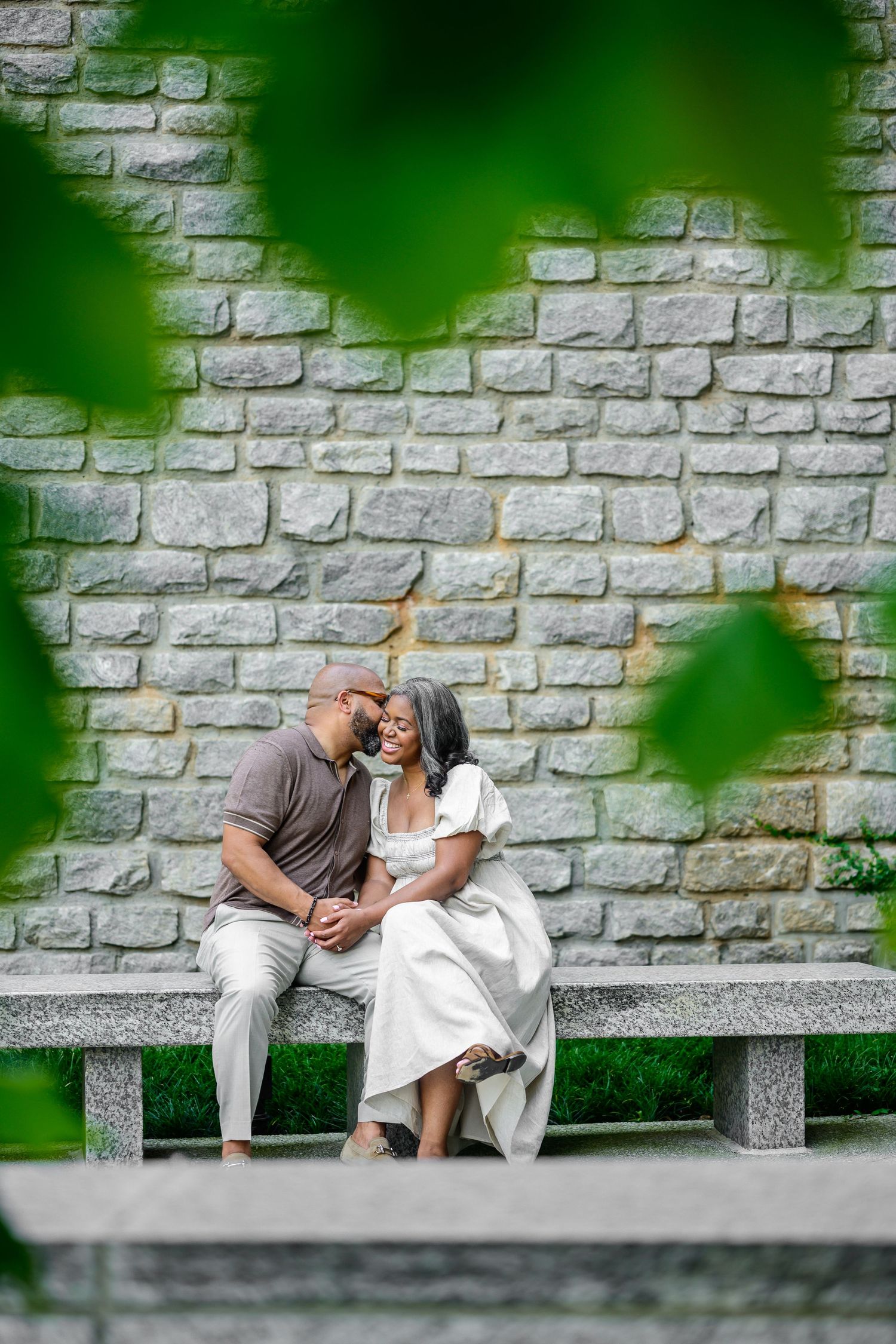 Stone wall and green foliage frame romantic couple's moment on concrete bench.