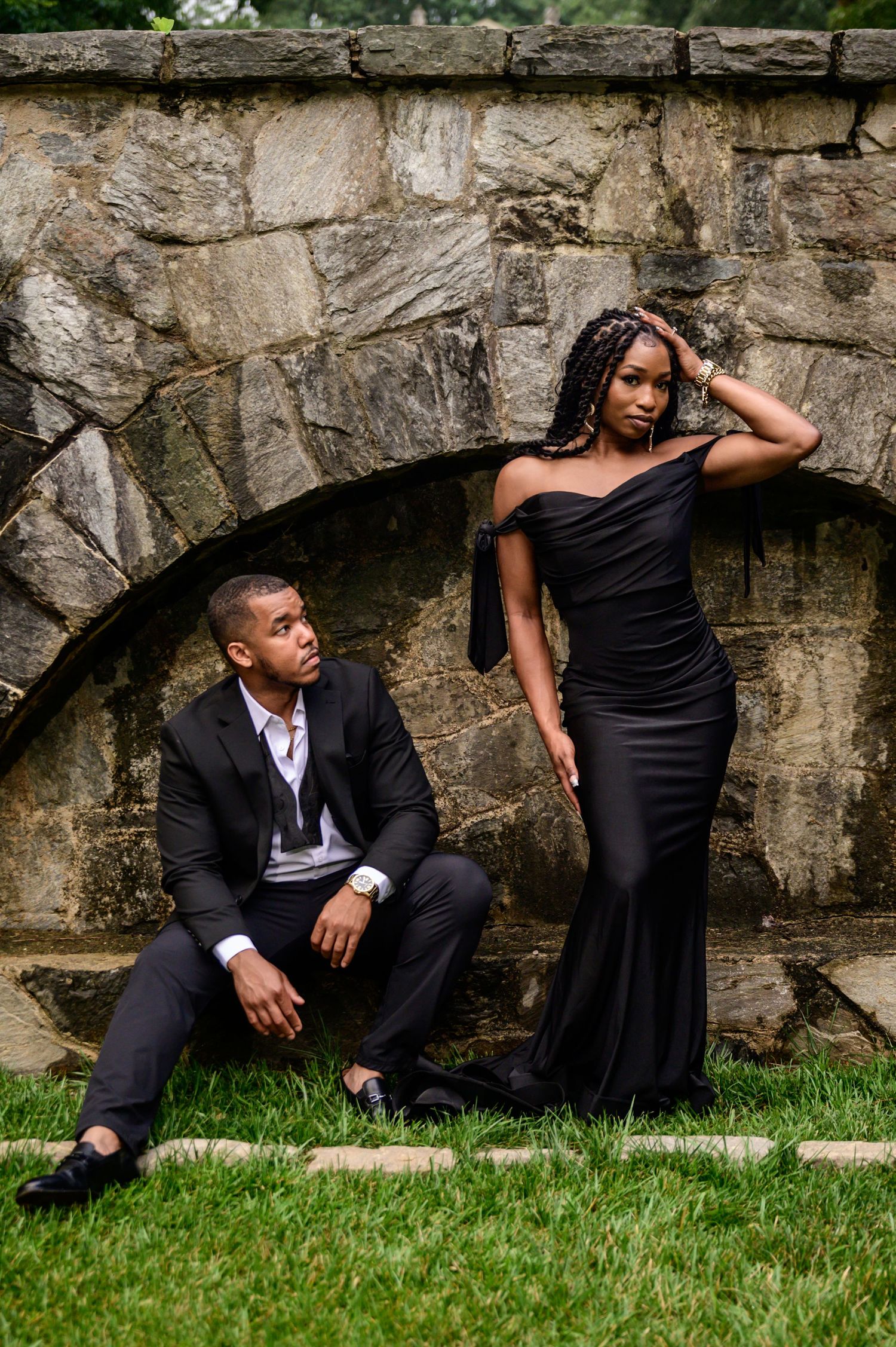 Couple in formal black attire poses artistically against stone wall and green grass.