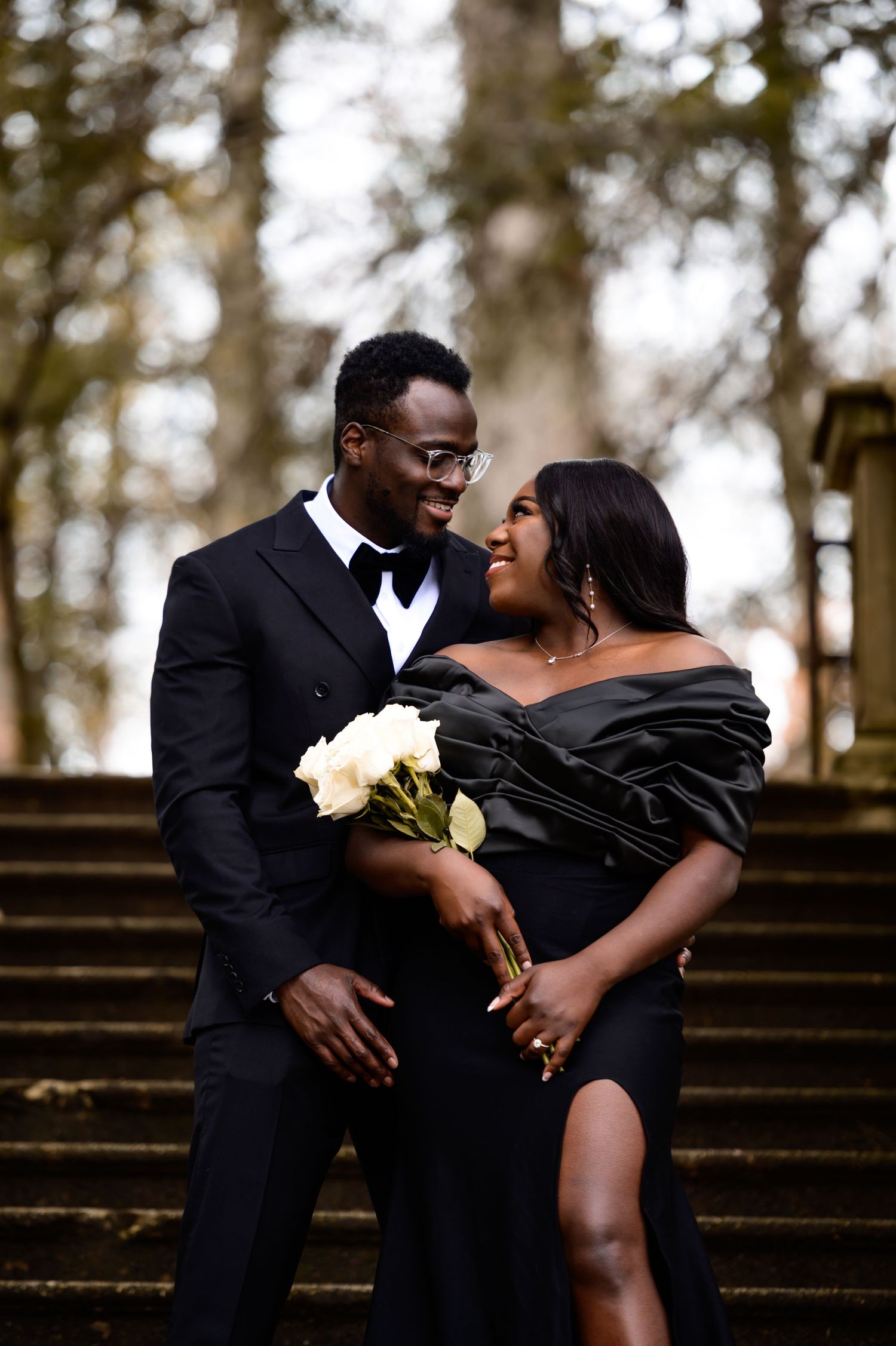 Joyful couple in formal black attire share loving moment on stone steps with white flowers.