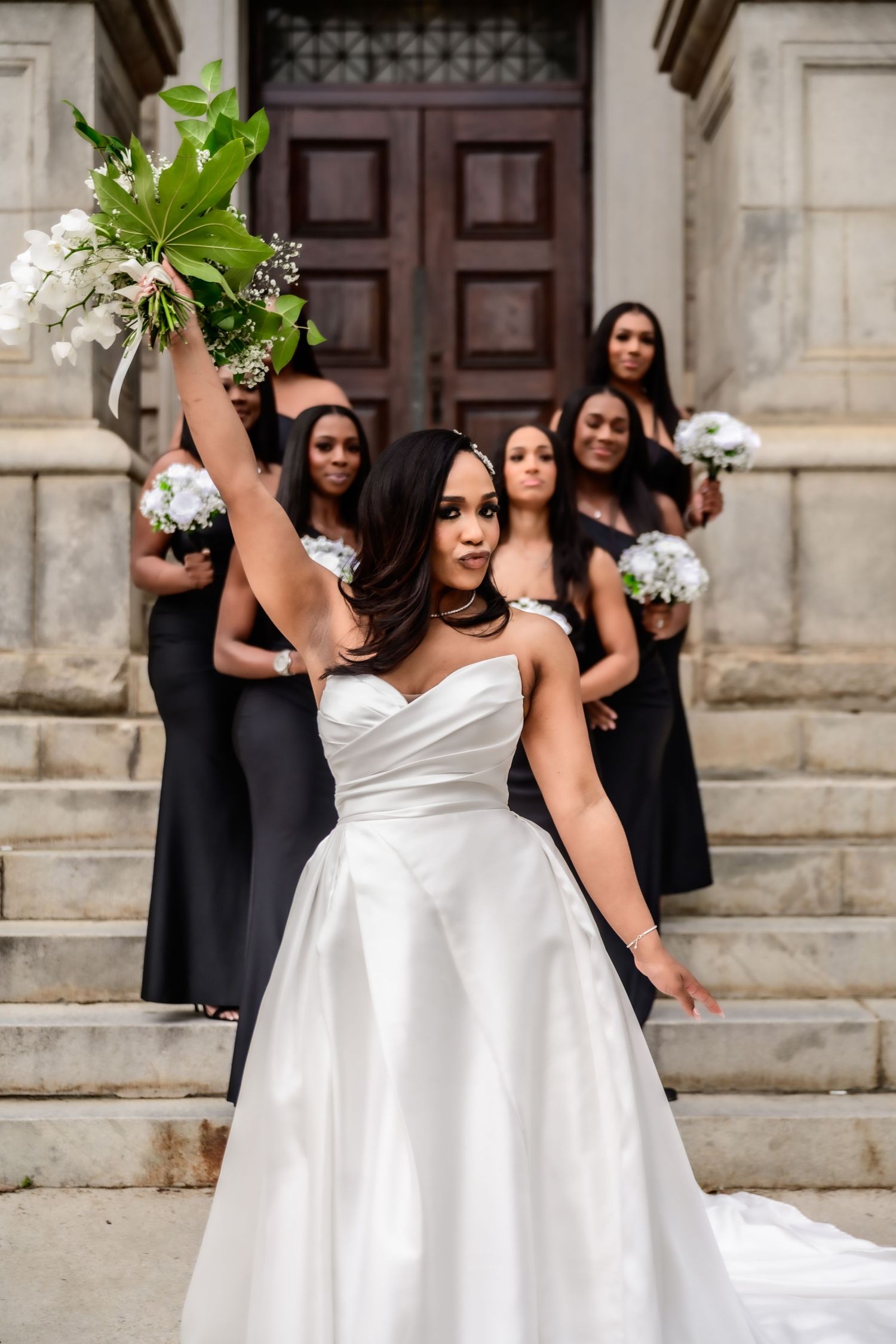 Bride in white dress celebrates with bridesmaids in black dresses on stone steps.
