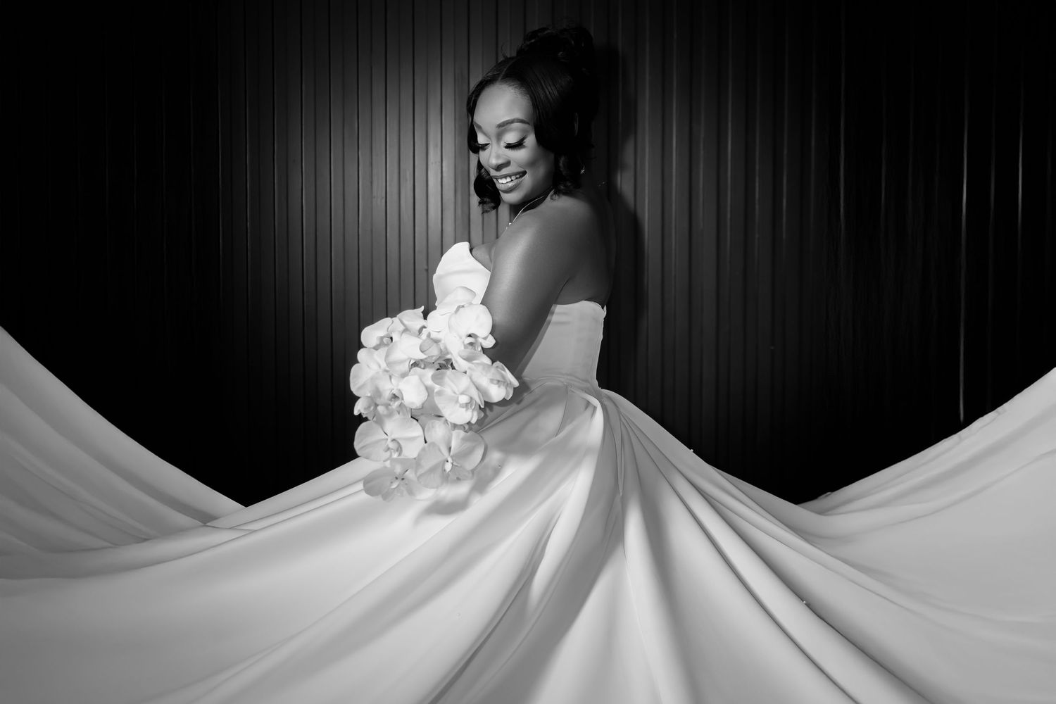 Black and white portrait of a bride in a flowing wedding gown holding a white bouquet against a dark background.