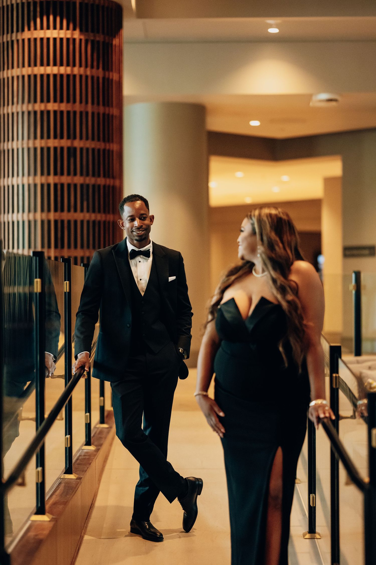 Elegant couple in formal black attire walking through a modern hotel hallway with architectural columns.