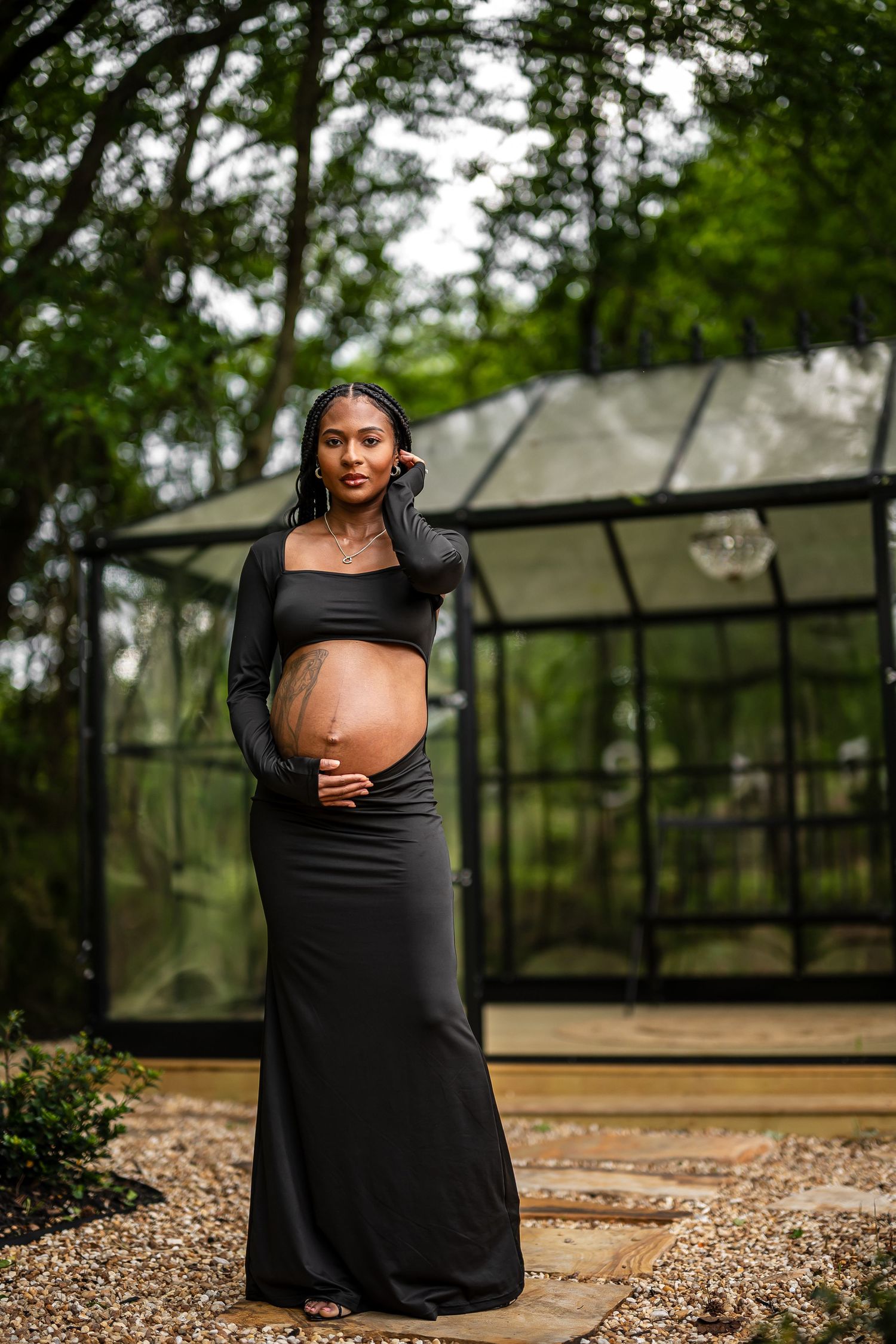 A maternity photoshoot in a black dress takes place in front of a modern greenhouse structure.