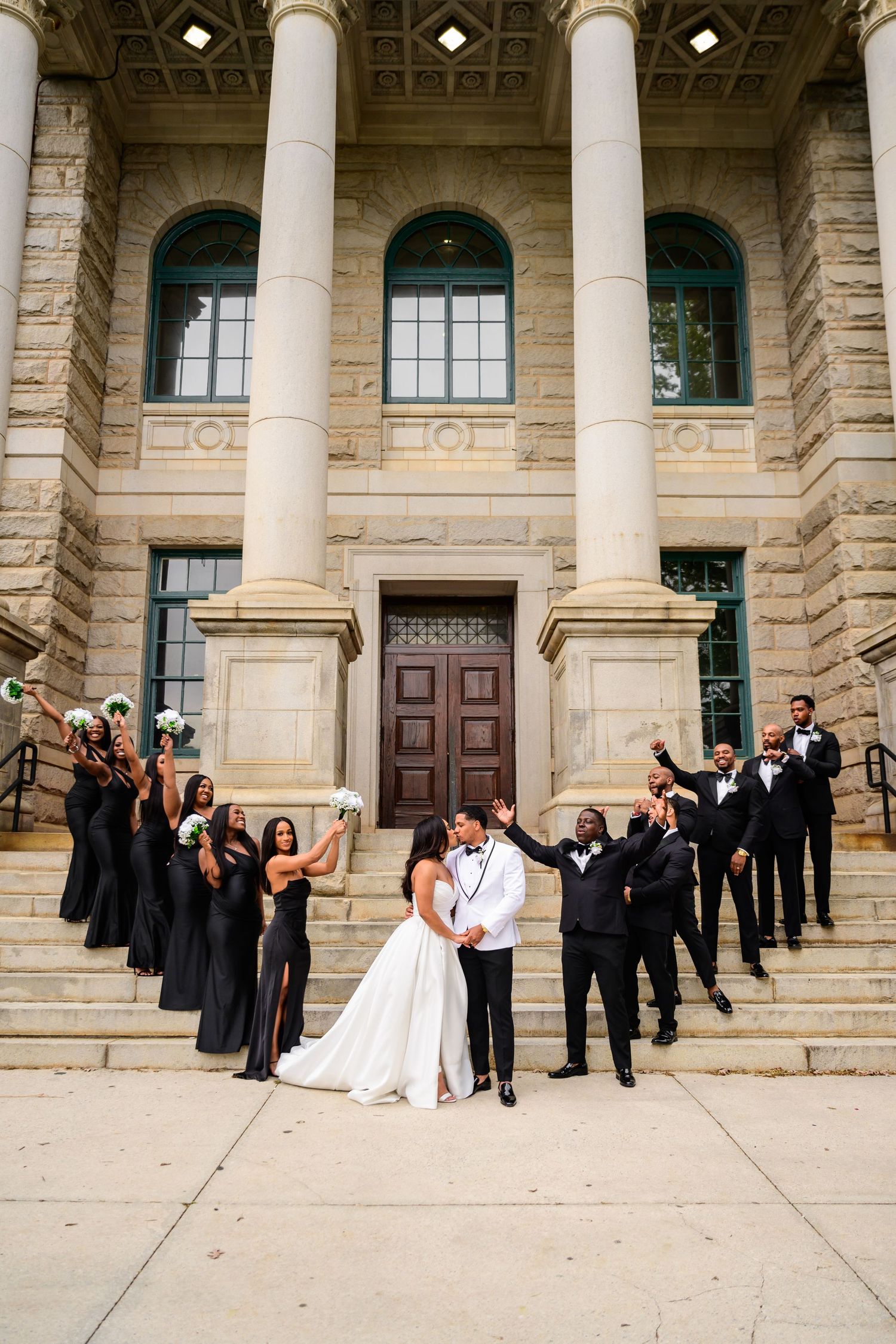 Wedding party celebrates on grand stone steps of classical building with white columns.