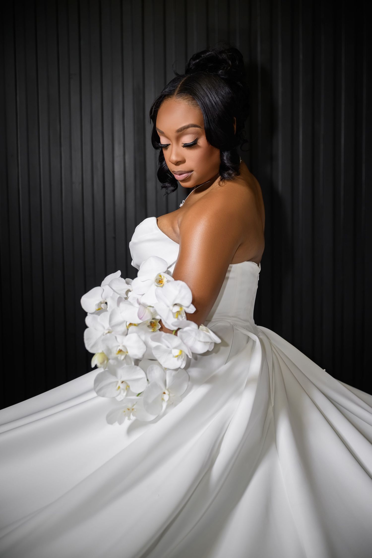 A stunning bridal portrait featuring an elegant white wedding gown with cascading orchids against a dark background.