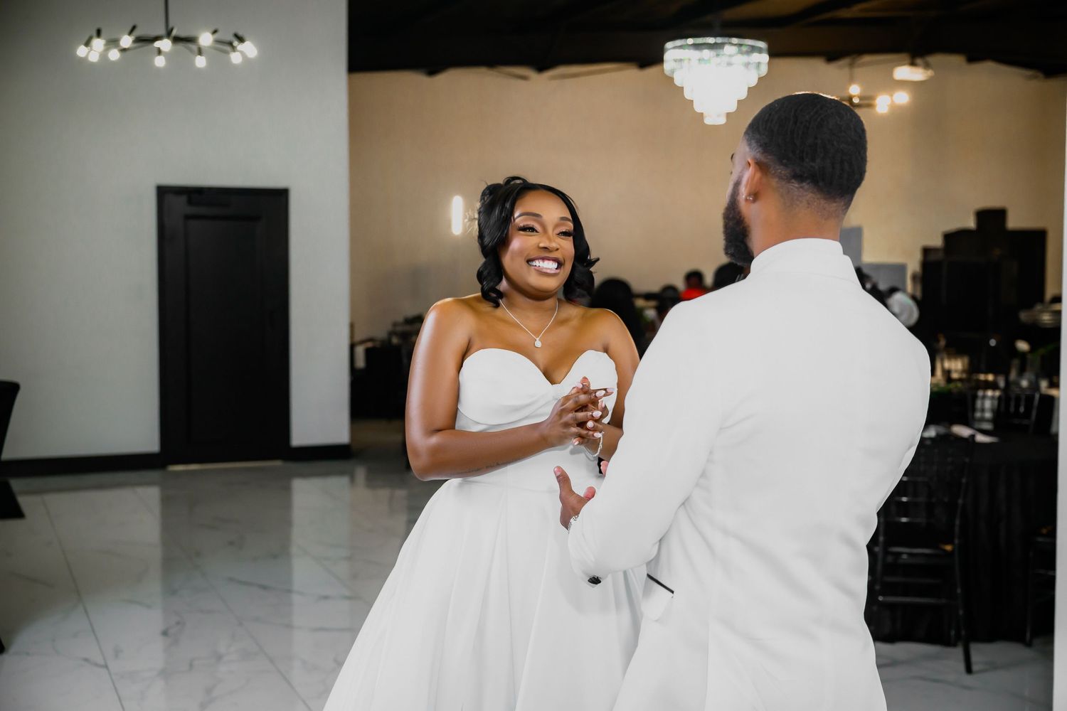 A couple shares their first dance in an elegant ballroom with chandeliers overhead during their wedding celebration.