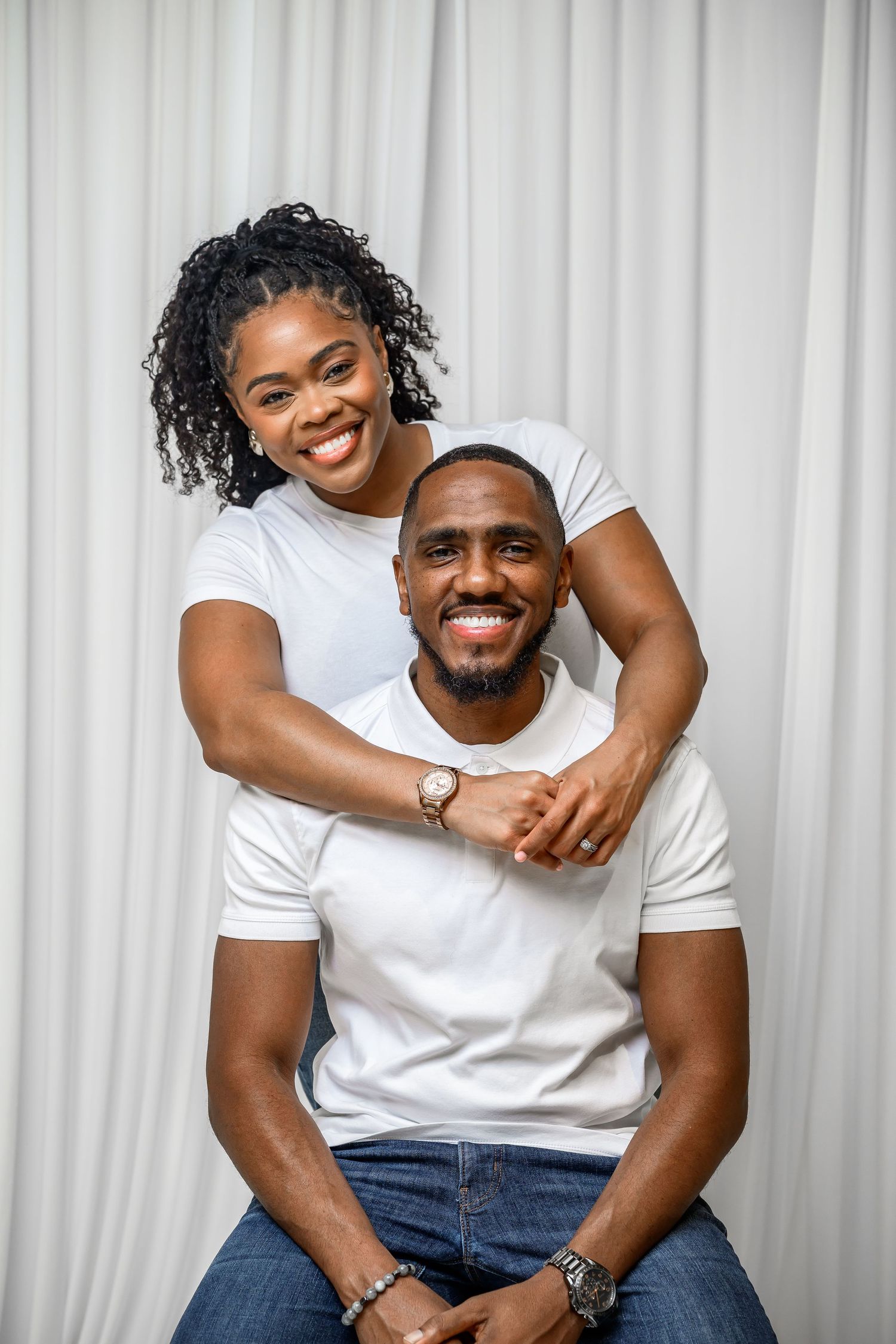 Couple wearing white t-shirts and jeans poses playfully against white curtain background in studio setting.