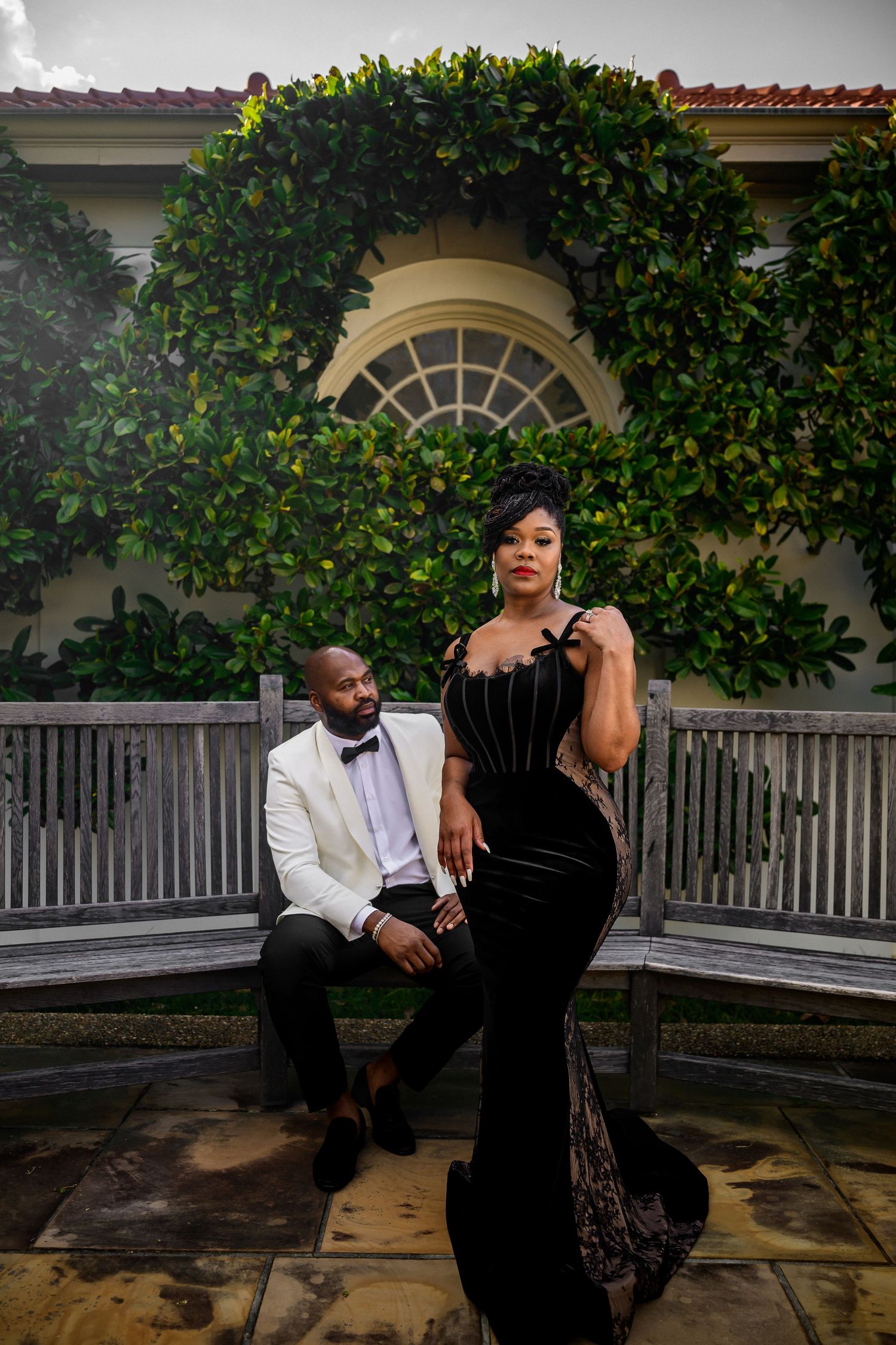 A couple poses together in front of lush green foliage and a white picket fence during an outdoor photo session.