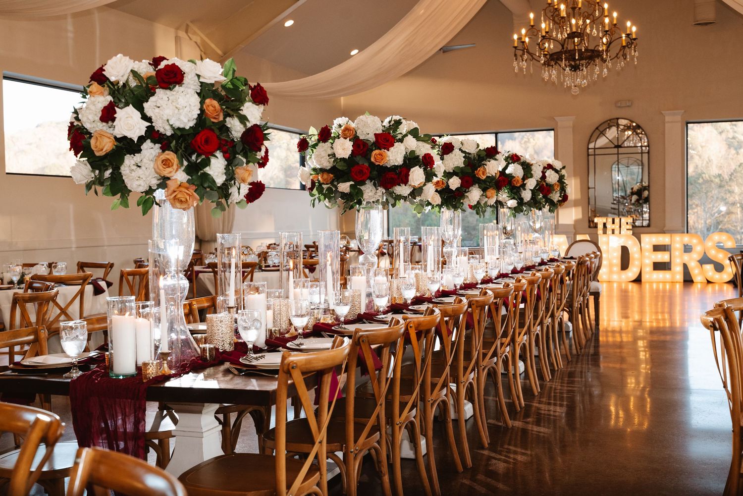 Long banquet tables with wooden crossback chairs arranged in an elegant reception venue with chandeliers.