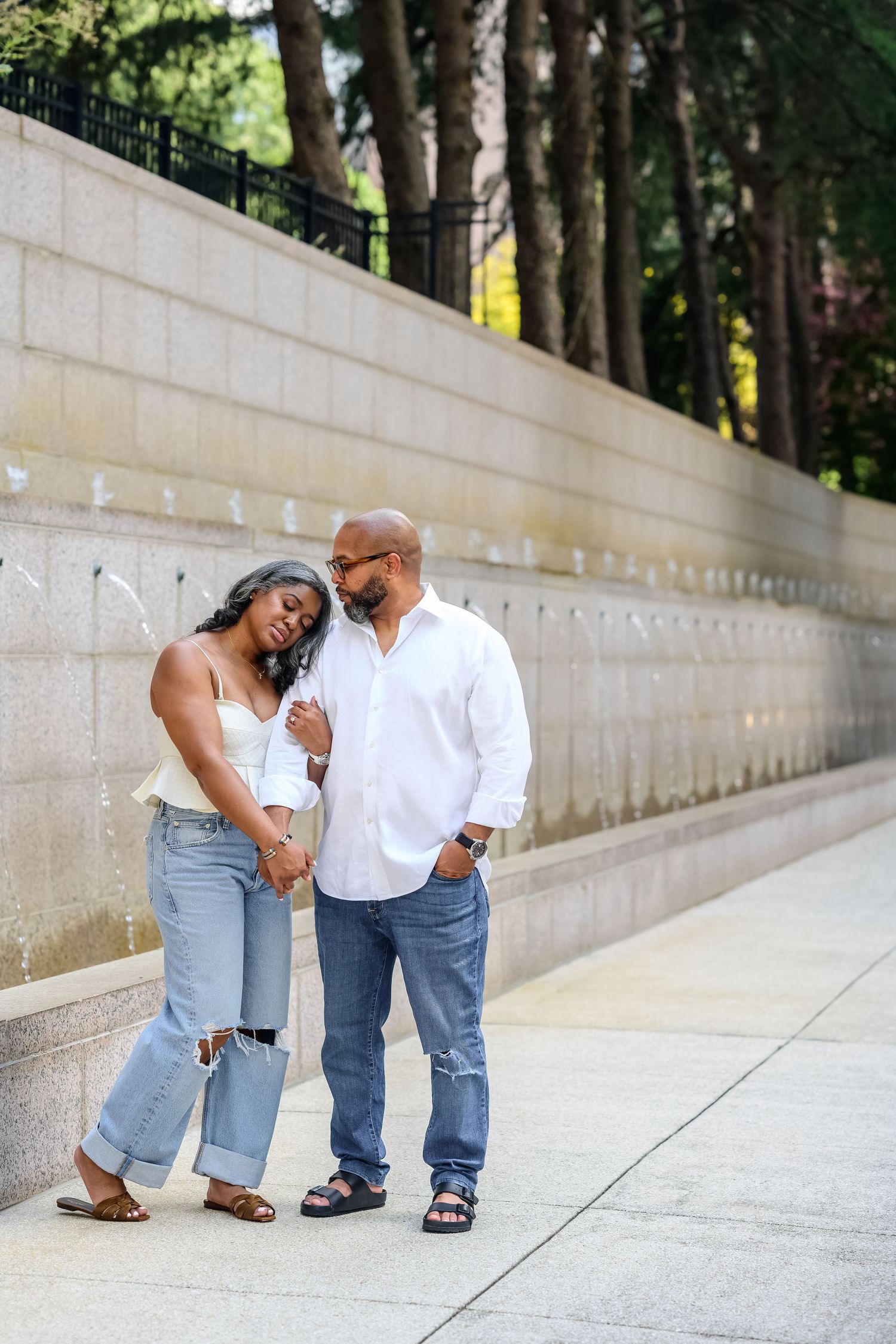 Couple in casual attire shares playful moment along concrete wall outdoors.