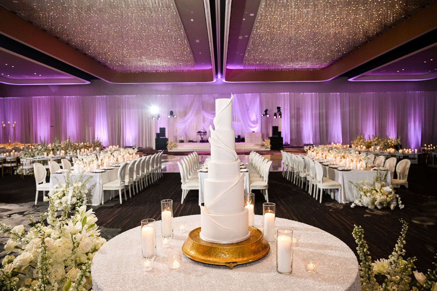 Tall white wedding cake displayed in center of elegant ballroom with purple ambient lighting.