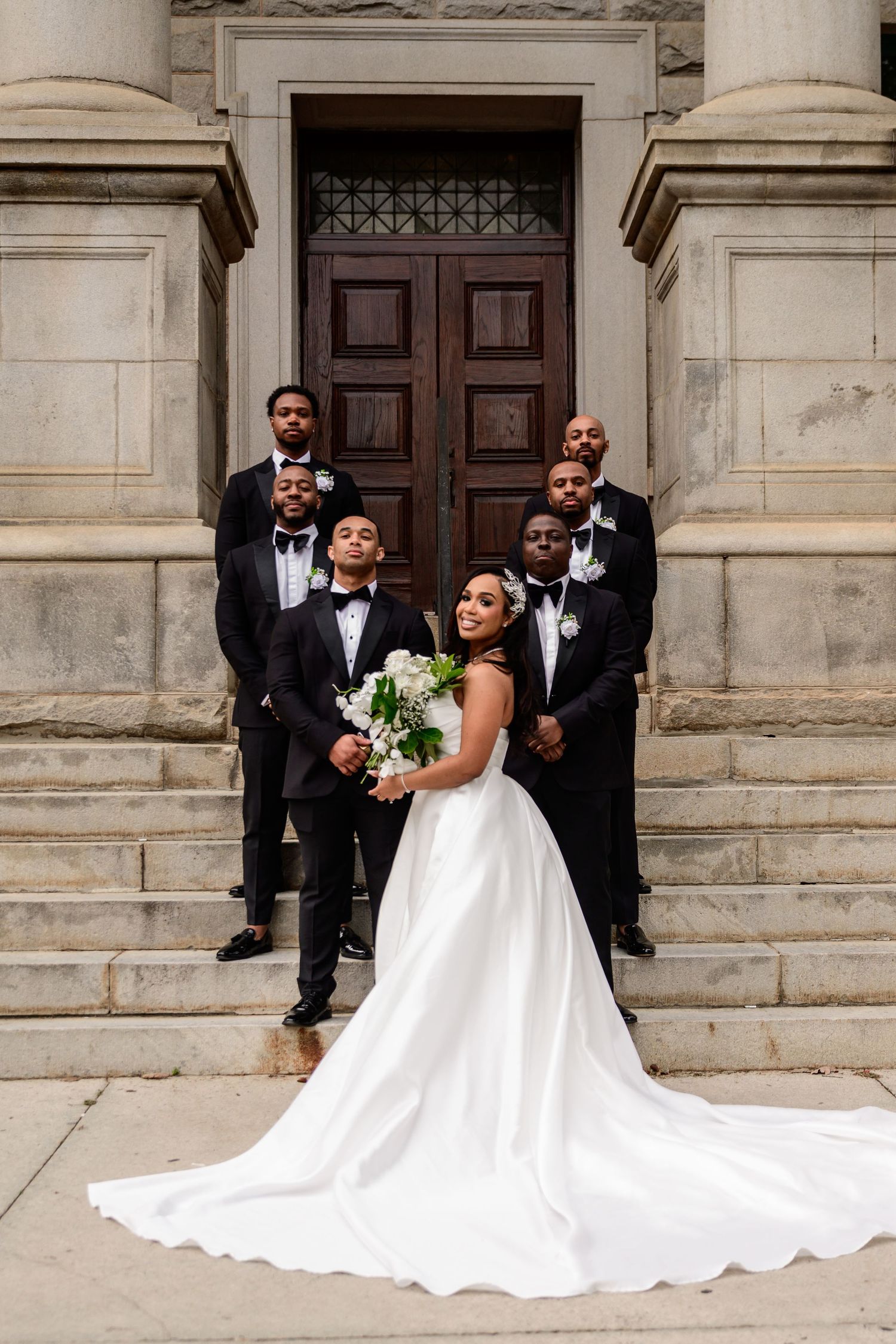 Bride in flowing white gown poses dramatically on stone steps with wedding party.